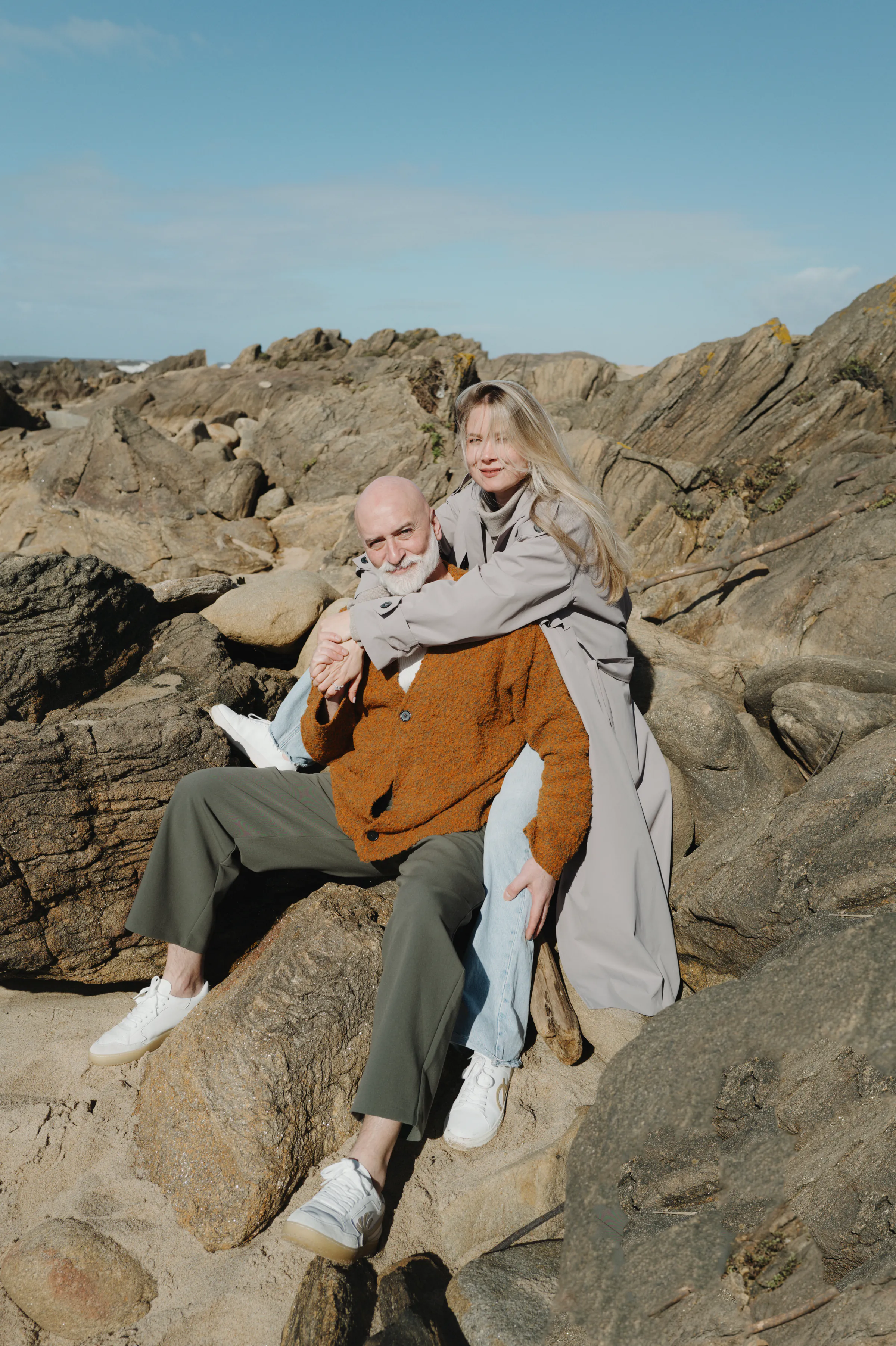Couple seated on sunlit seaside rocks during a relaxed outdoor portrait session.