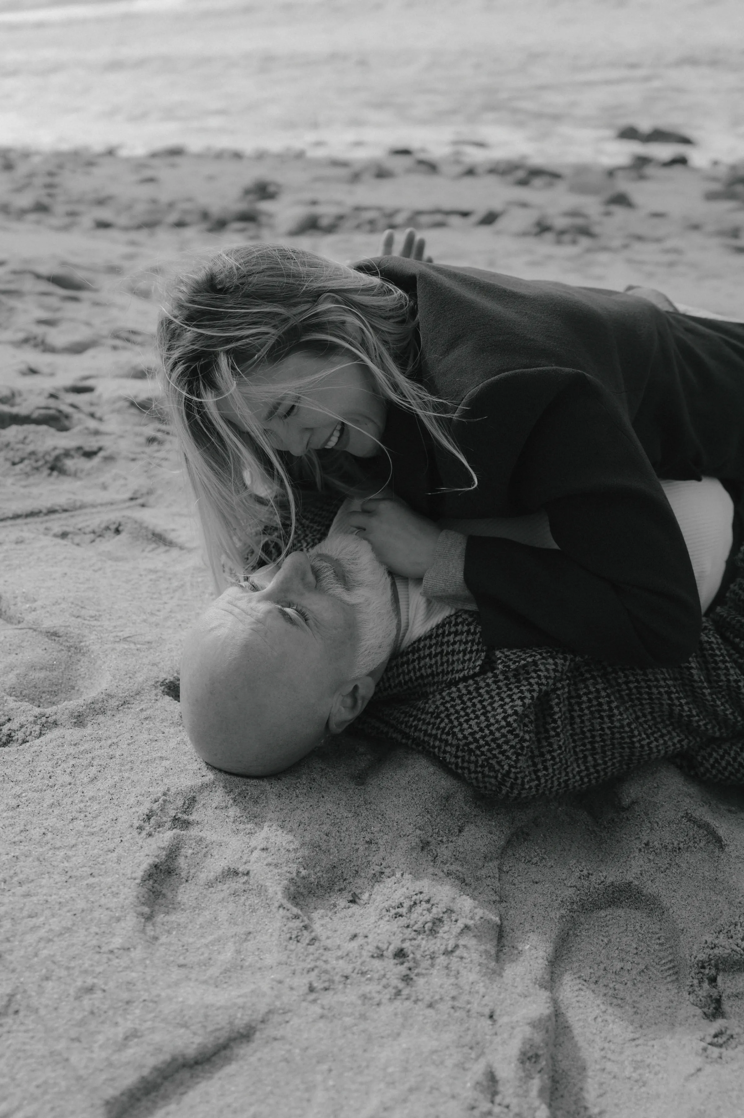 Black-and-white seaside portrait of a couple lying close together on sandy beach during an outdoor session.