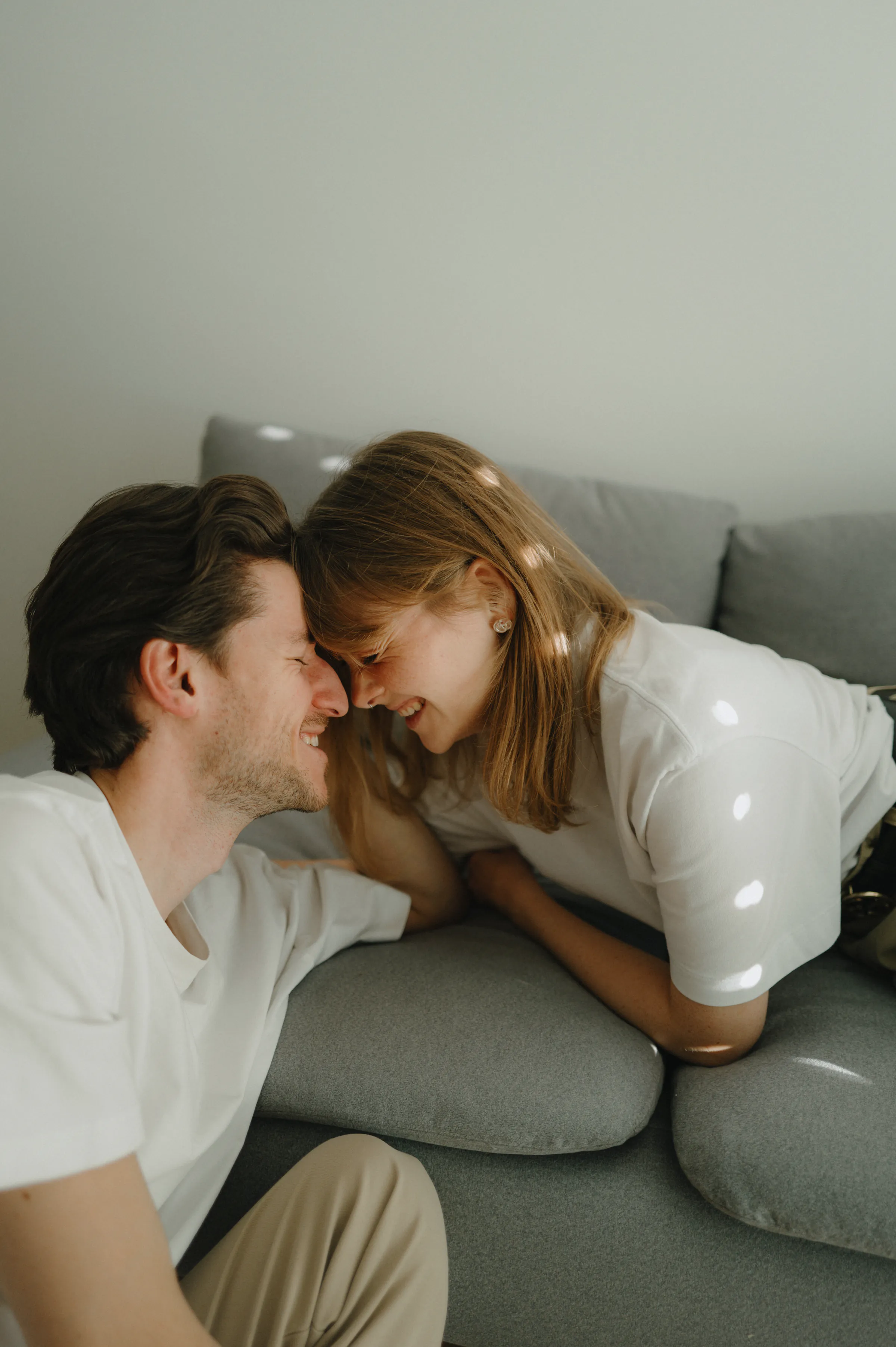 Couple leaning forehead to forehead on a gray sofa during a soft light home session.