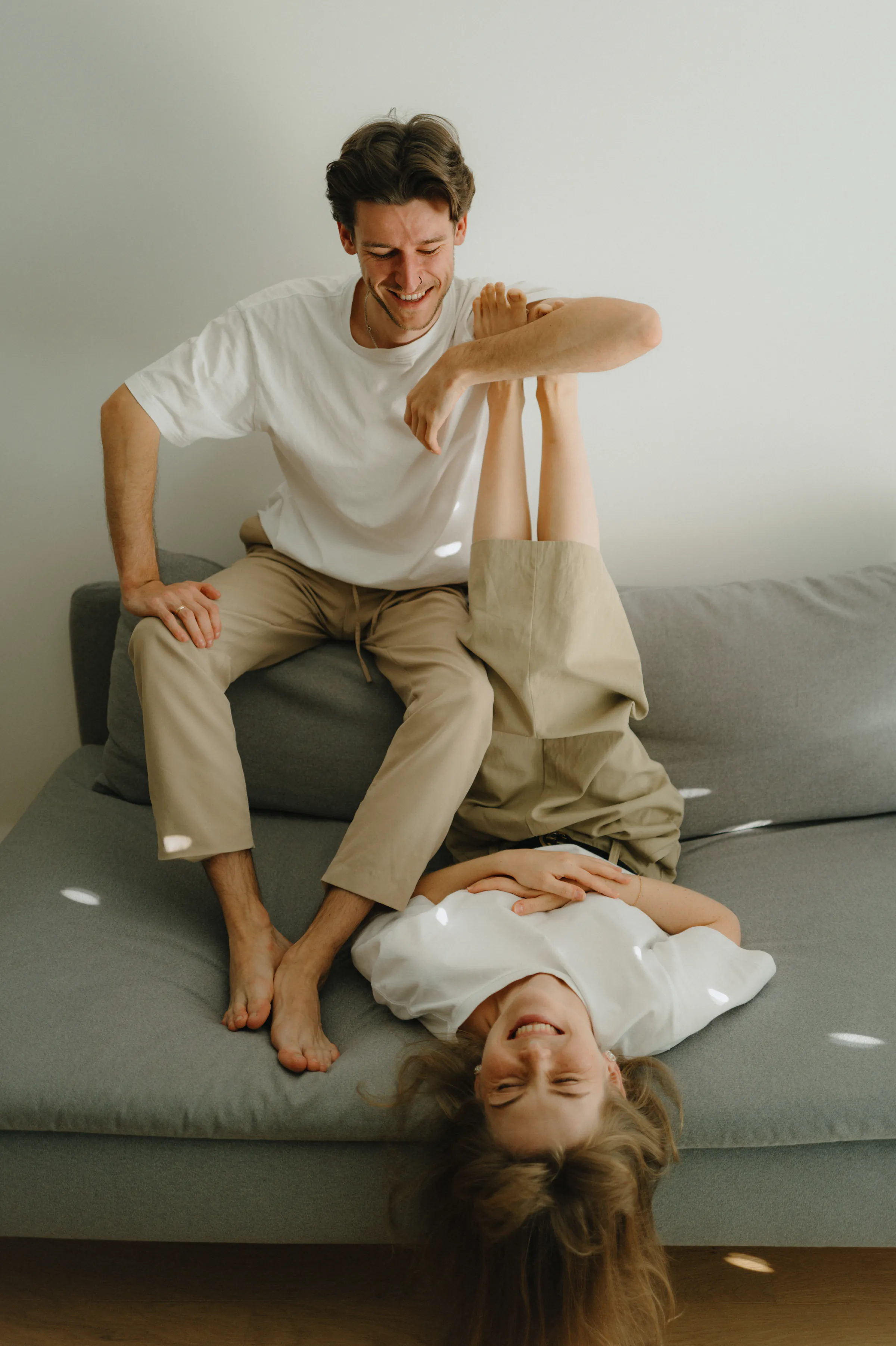 Couple during a relaxed home session on a gray sofa, one lounging upside down in soft light.