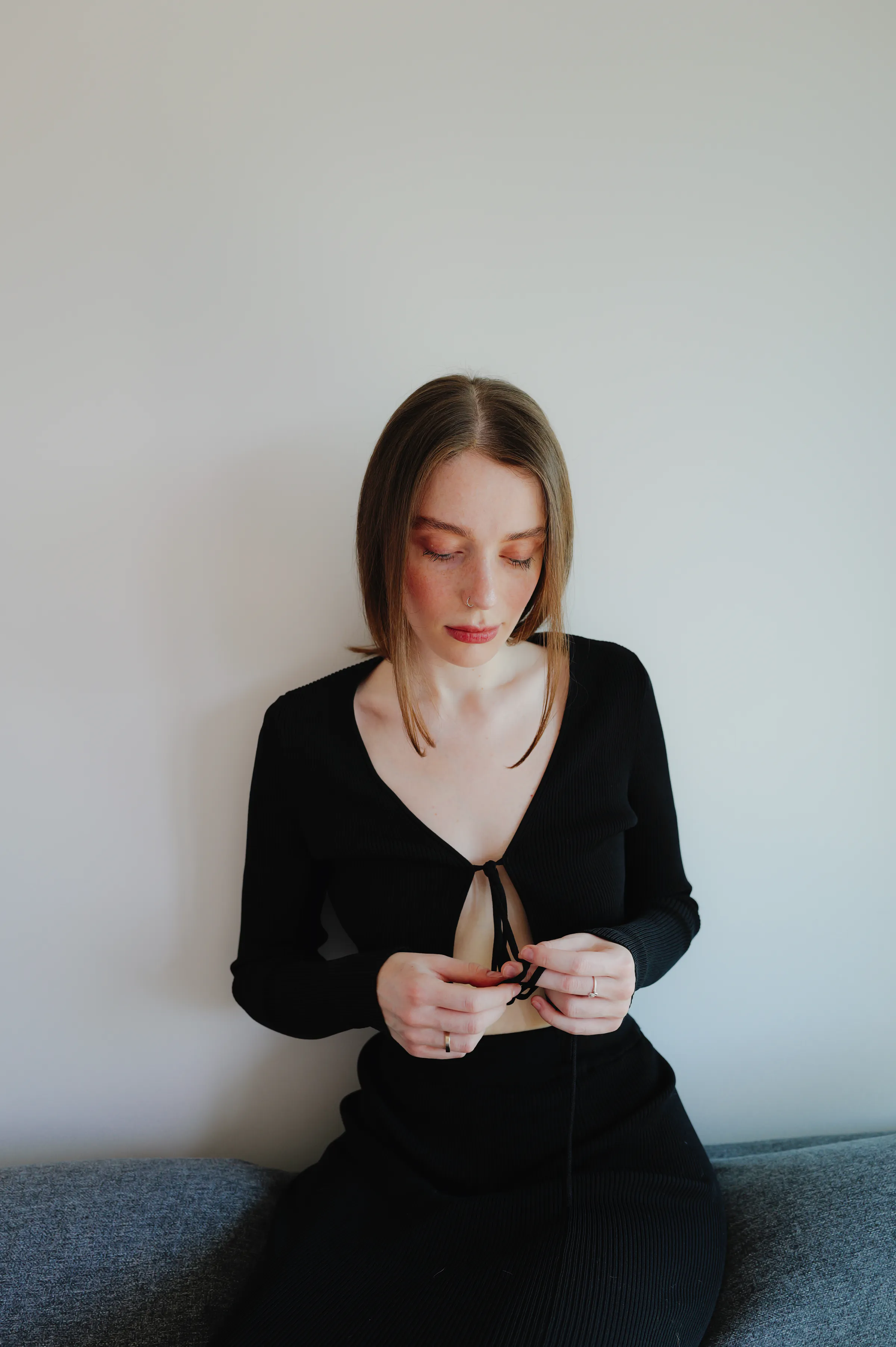 Indoor portrait of an adult seated on a sofa in soft light, looking down against a plain wall.