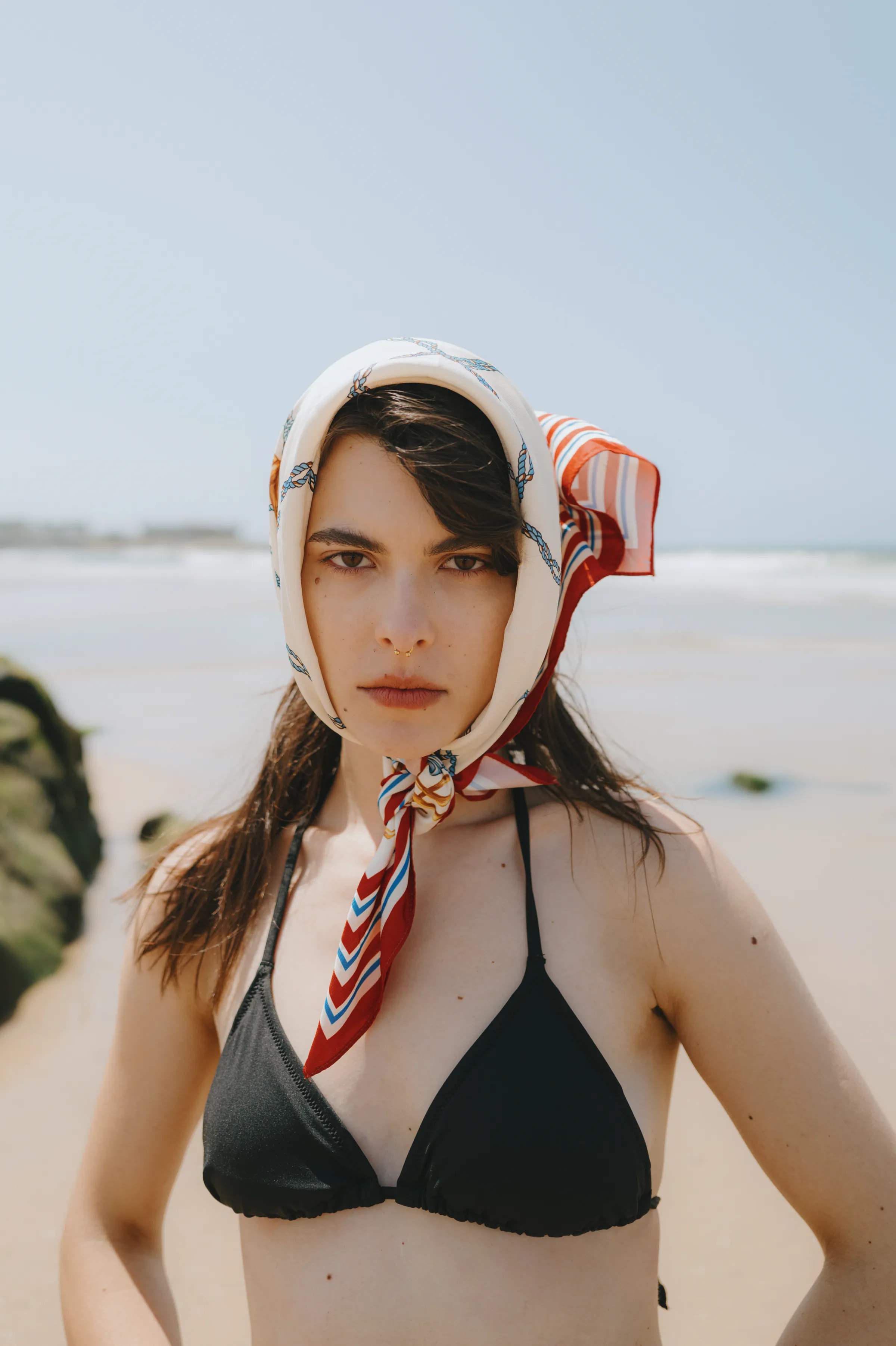 Close seaside portrait of a woman in a black bikini and patterned headscarf under soft daylight.