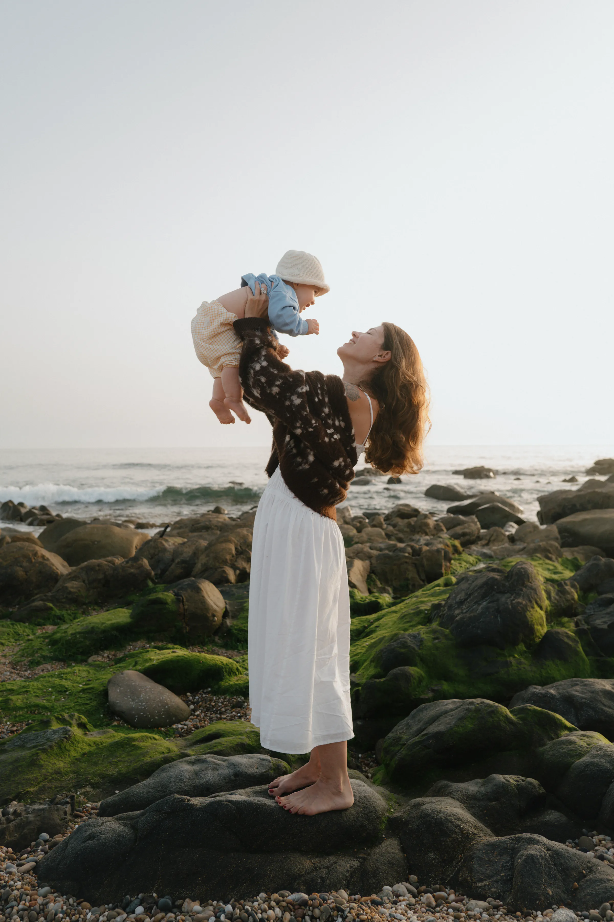 Woman lifting a baby during a soft light seaside session on a rocky shore.