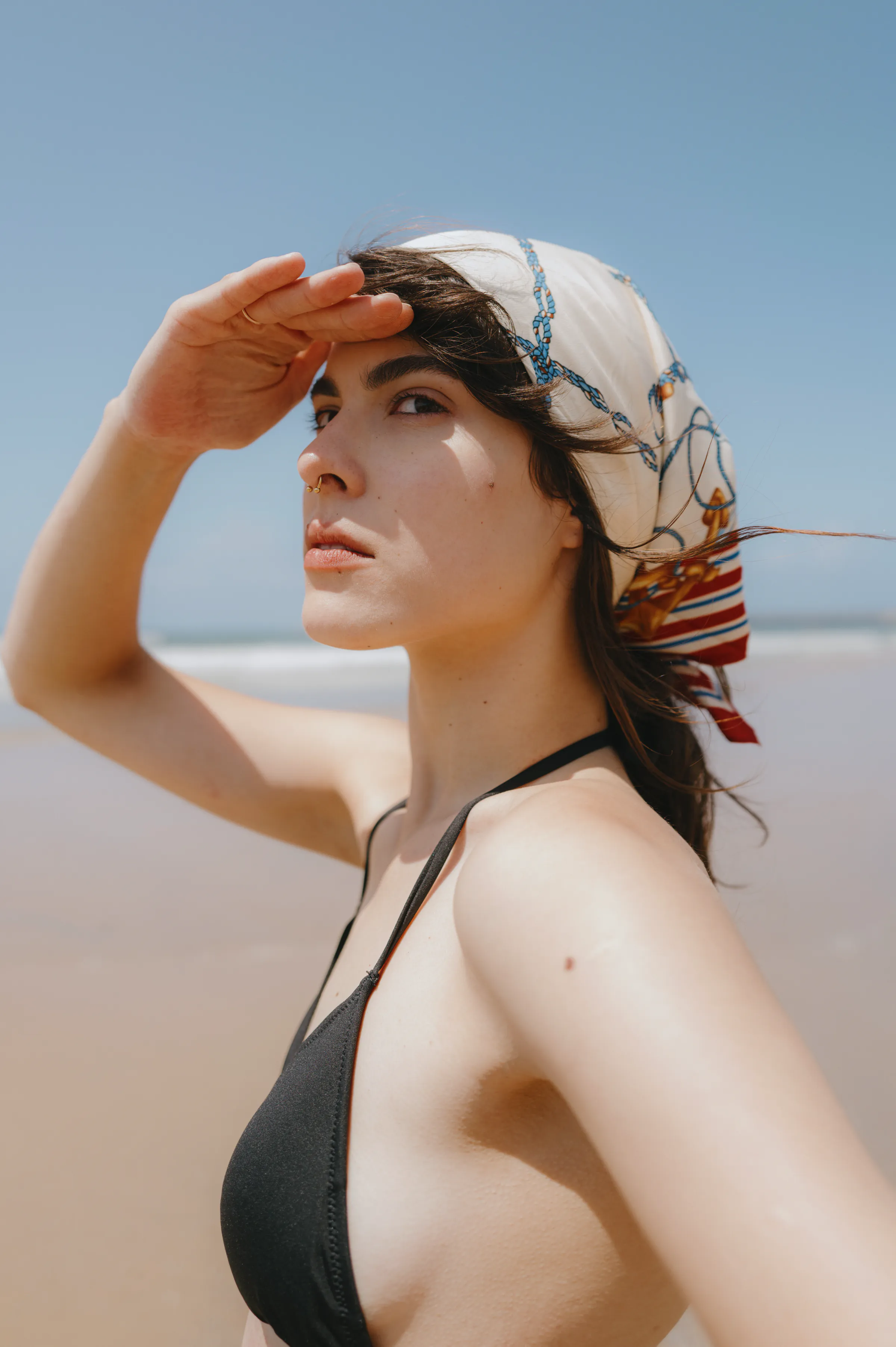 Seaside portrait of a person in a headscarf and black swimsuit, shading their eyes.
