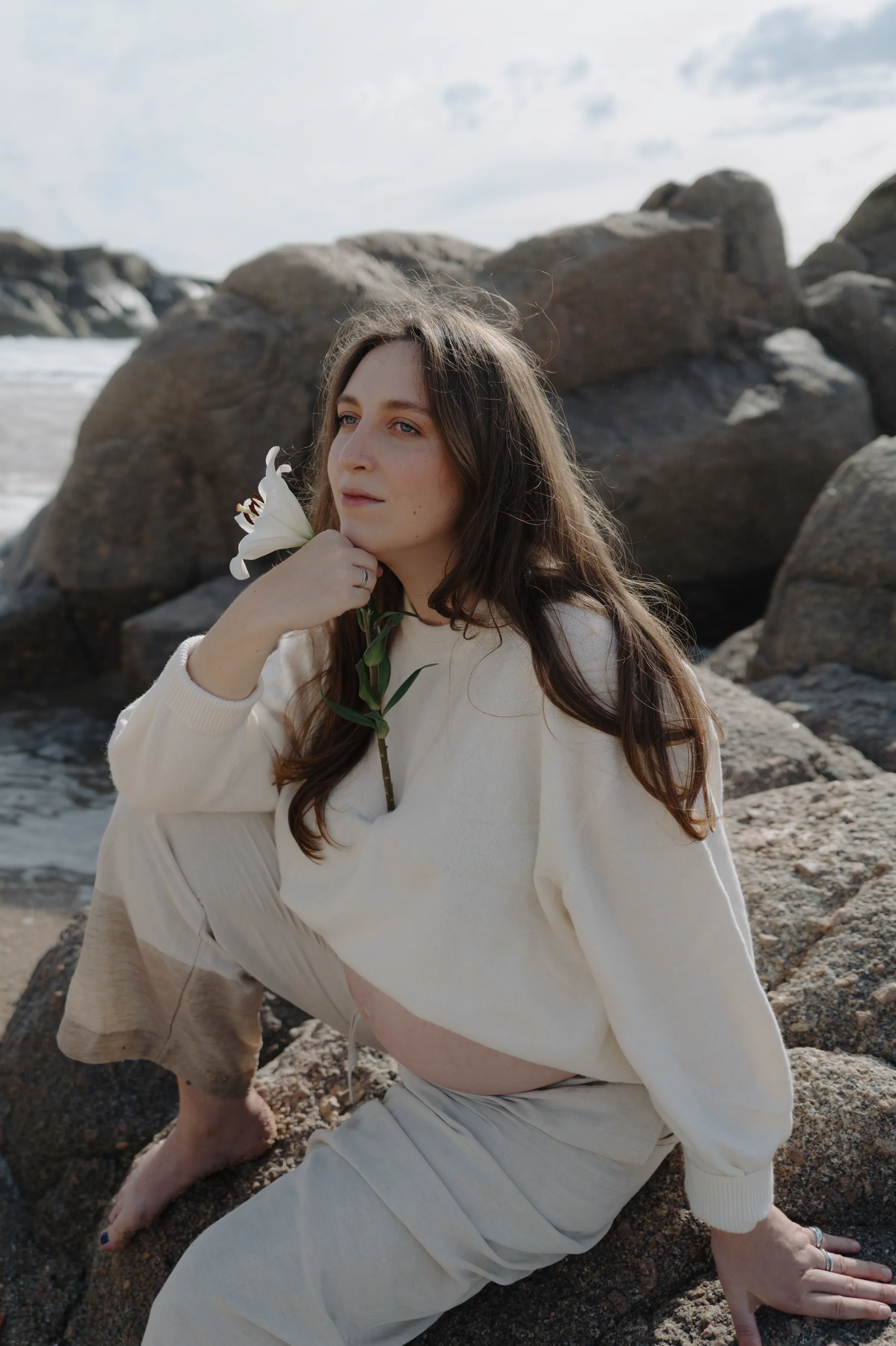 Seaside portrait of a woman seated on rocks, holding a white flower in soft light.