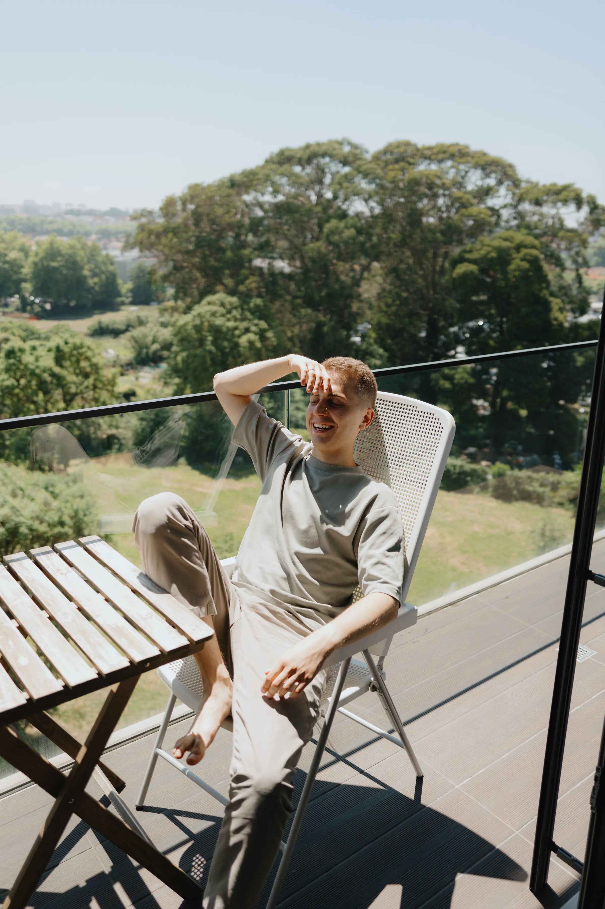 Outdoor portrait of an adult relaxing on a balcony chair in soft light, with trees beyond.