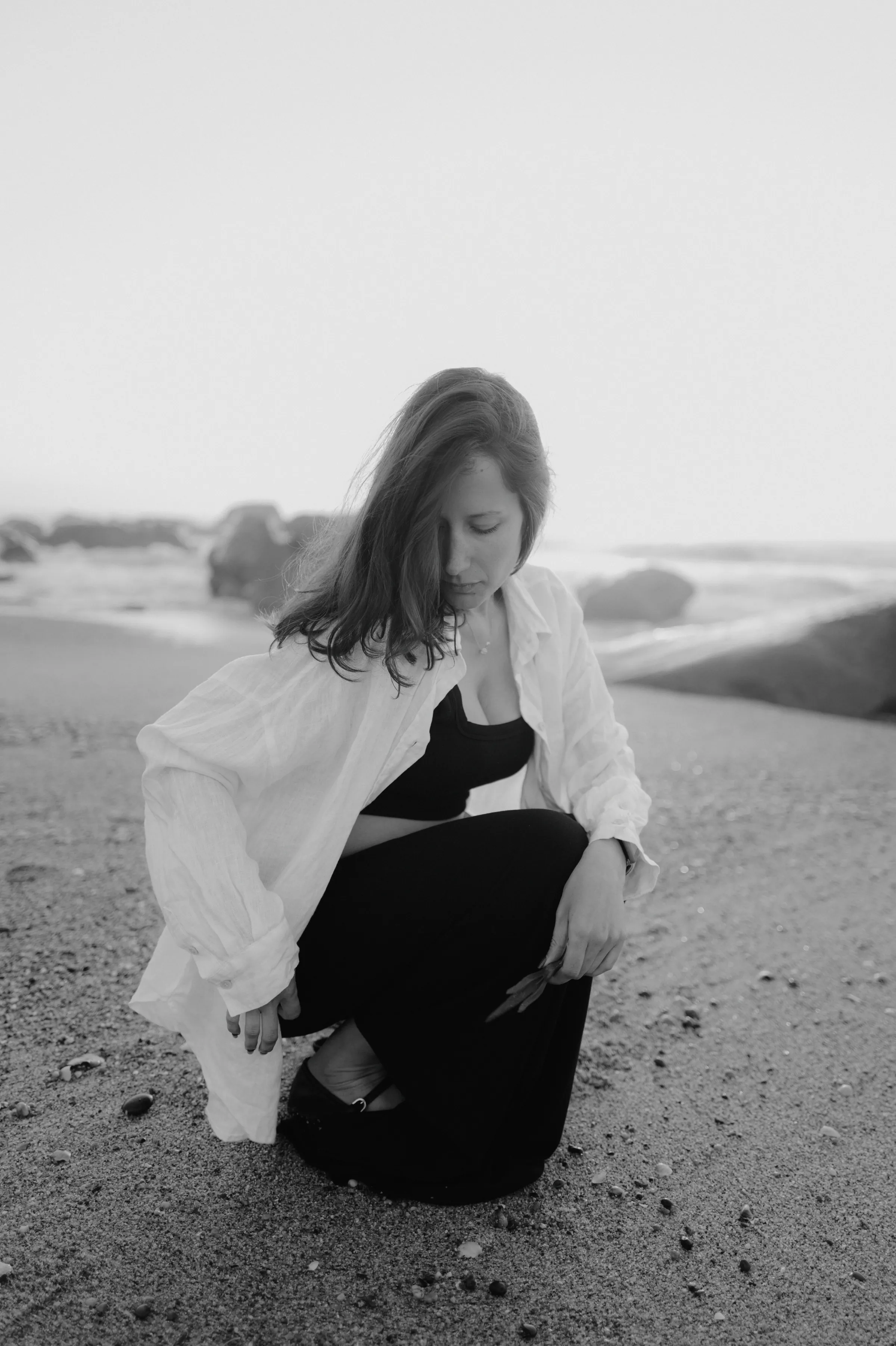 Black-and-white seaside portrait of an adult seated on sand in soft light, with rocks and surf behind.