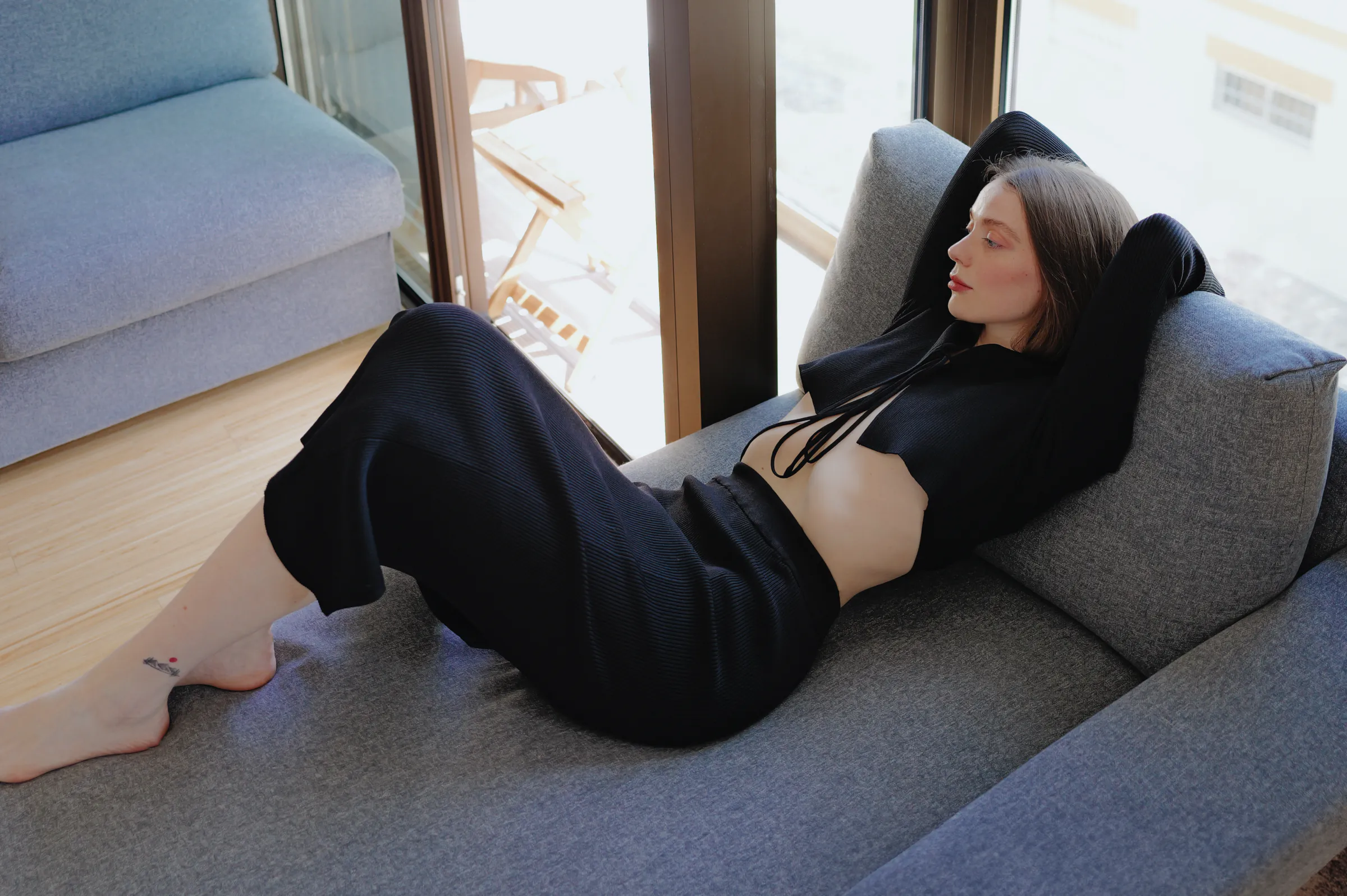 Indoor portrait of a person reclining on a gray sofa in soft window light.