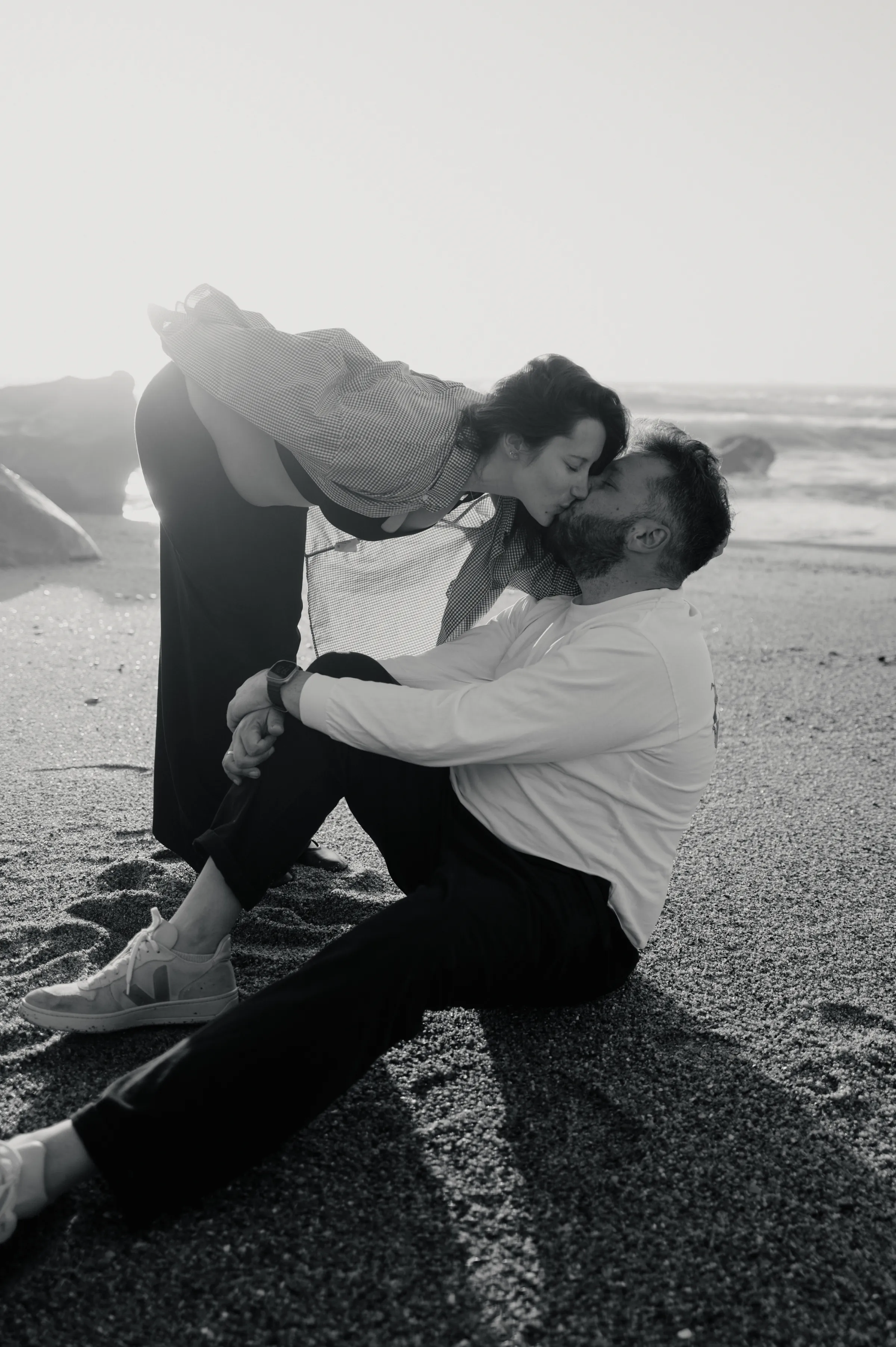 Black-and-white seaside portrait of a couple kissing on a pebbled beach in soft light.