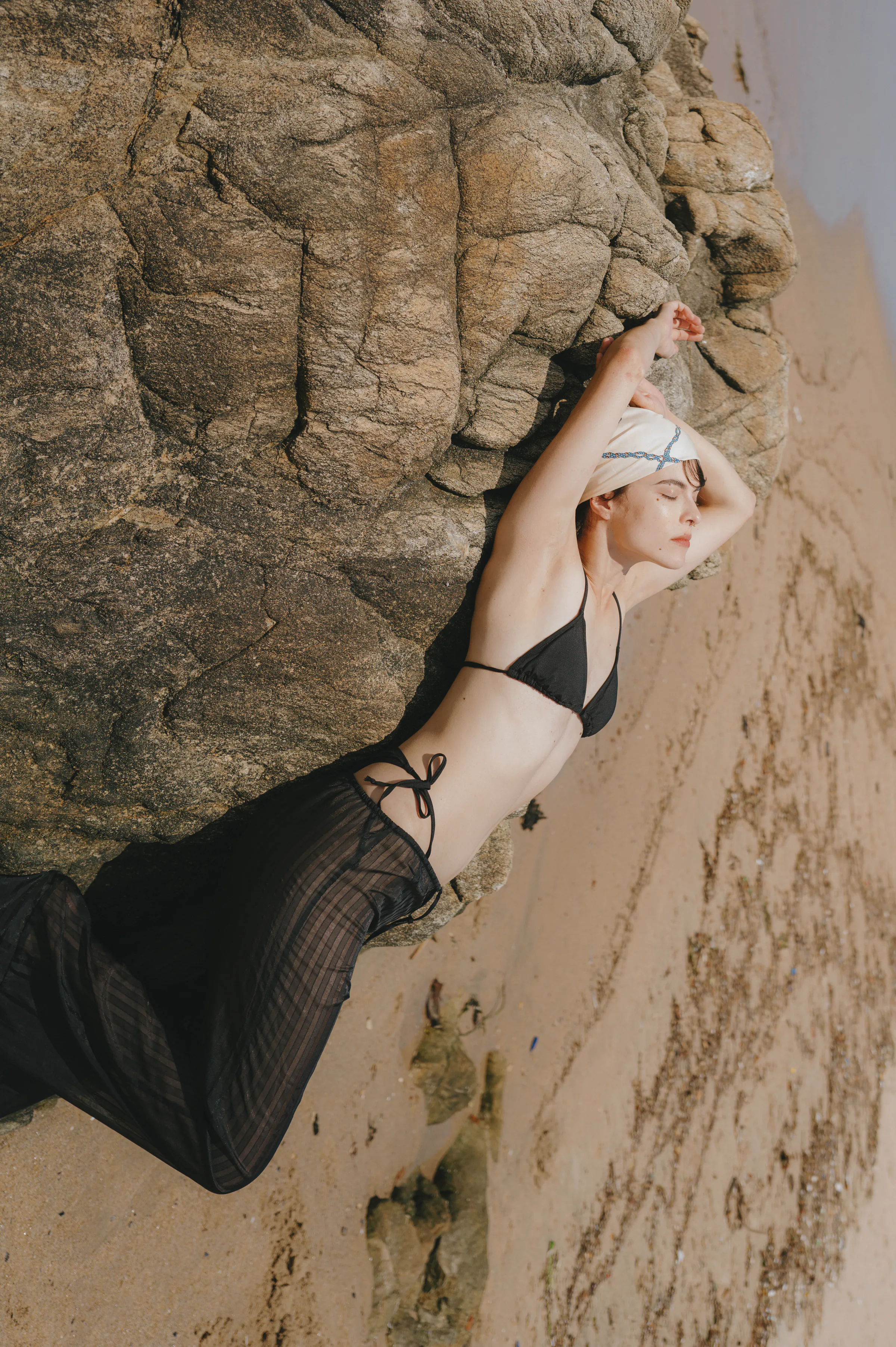Outdoor seaside portrait of a person reclining against sunlit rocks in a black swim top and sheer skirt