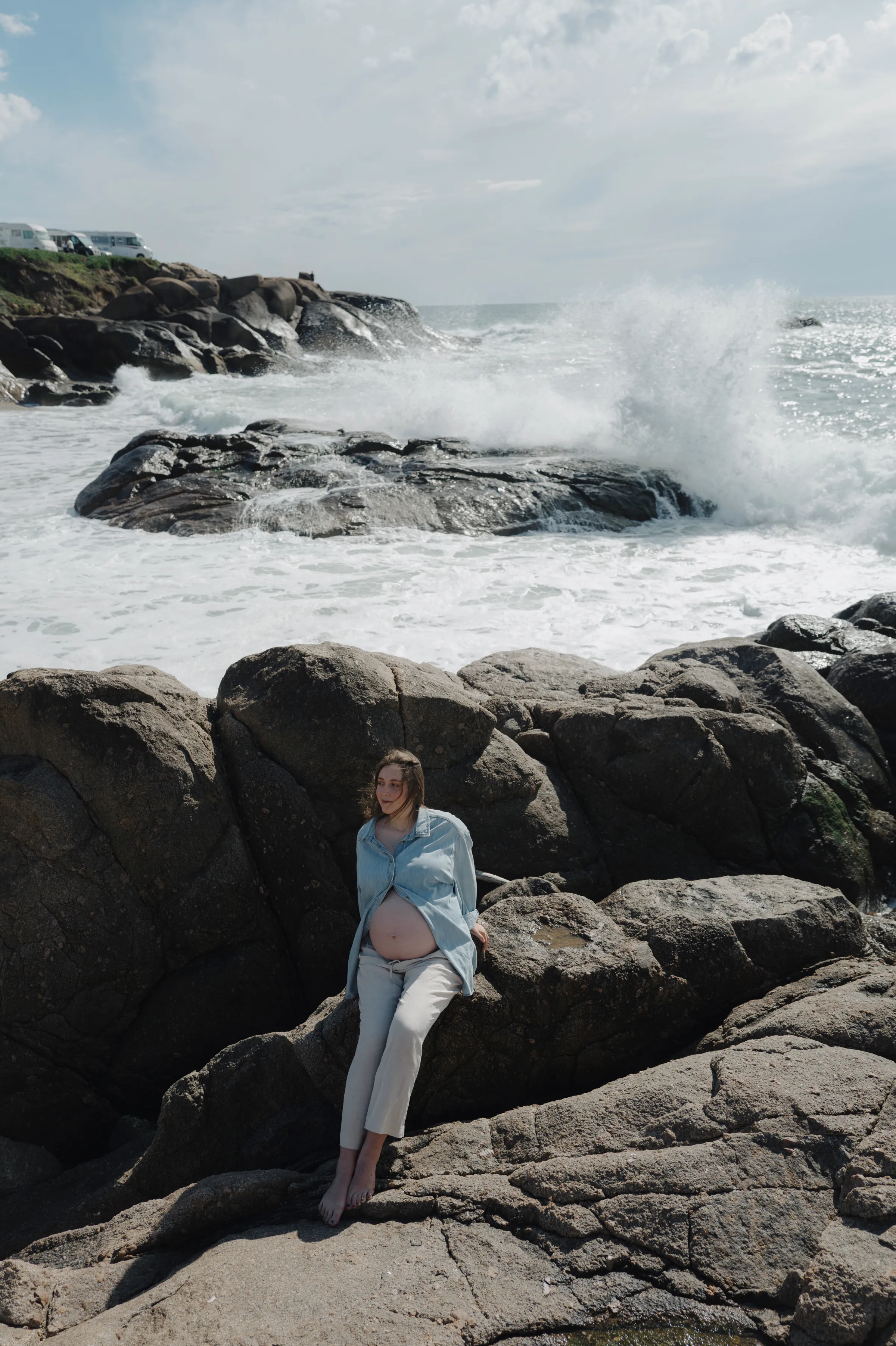 Pregnancy portrait on seaside rocks with waves breaking behind in soft daylight