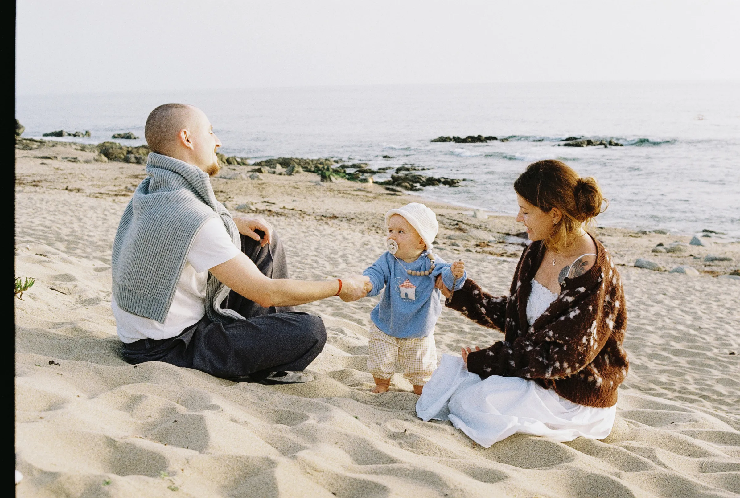 Family seaside session with two adults and a baby sitting on sand near calm water.