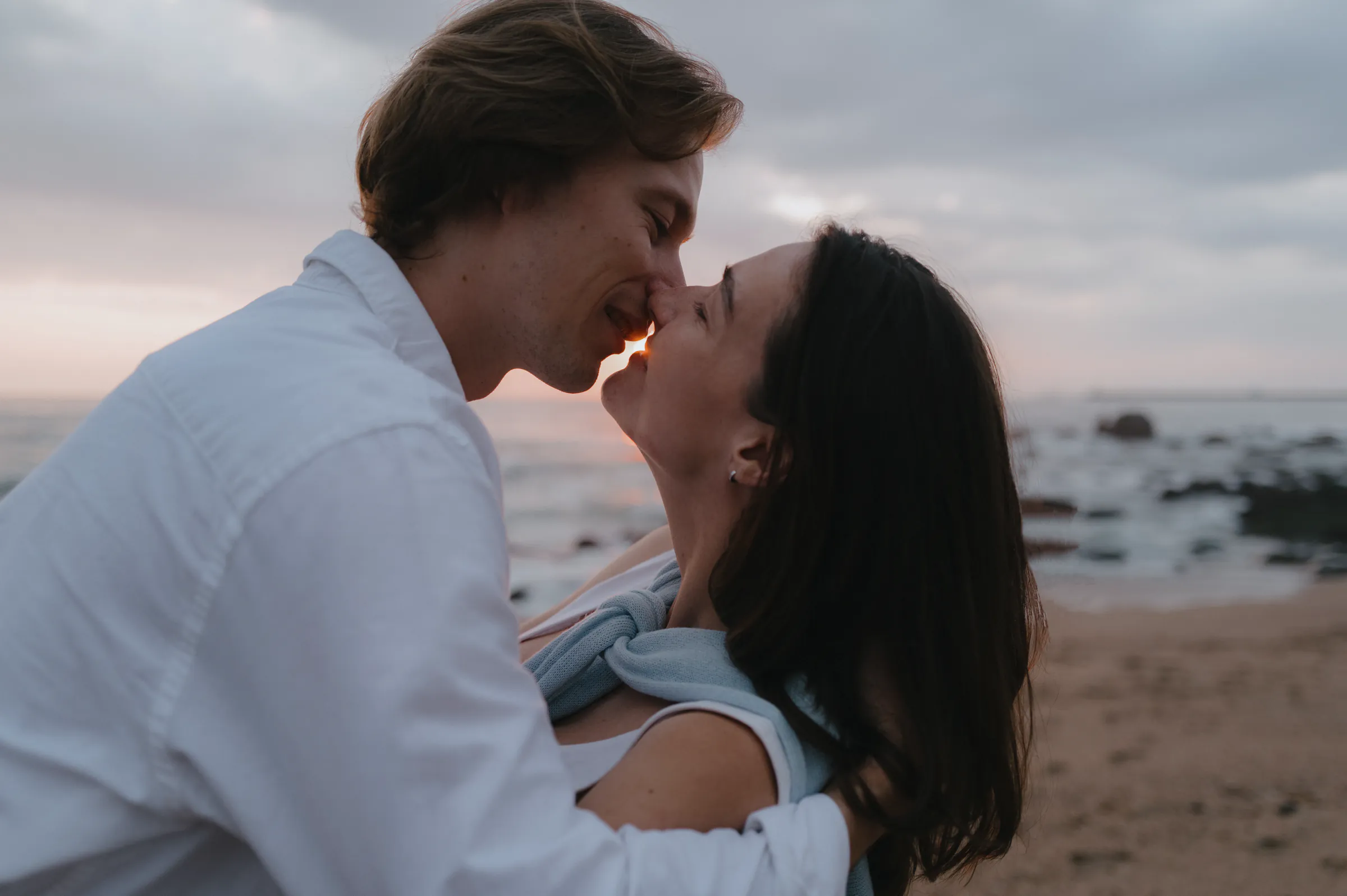 Couple in a close seaside portrait, leaning together on a cloudy beach at dusk.