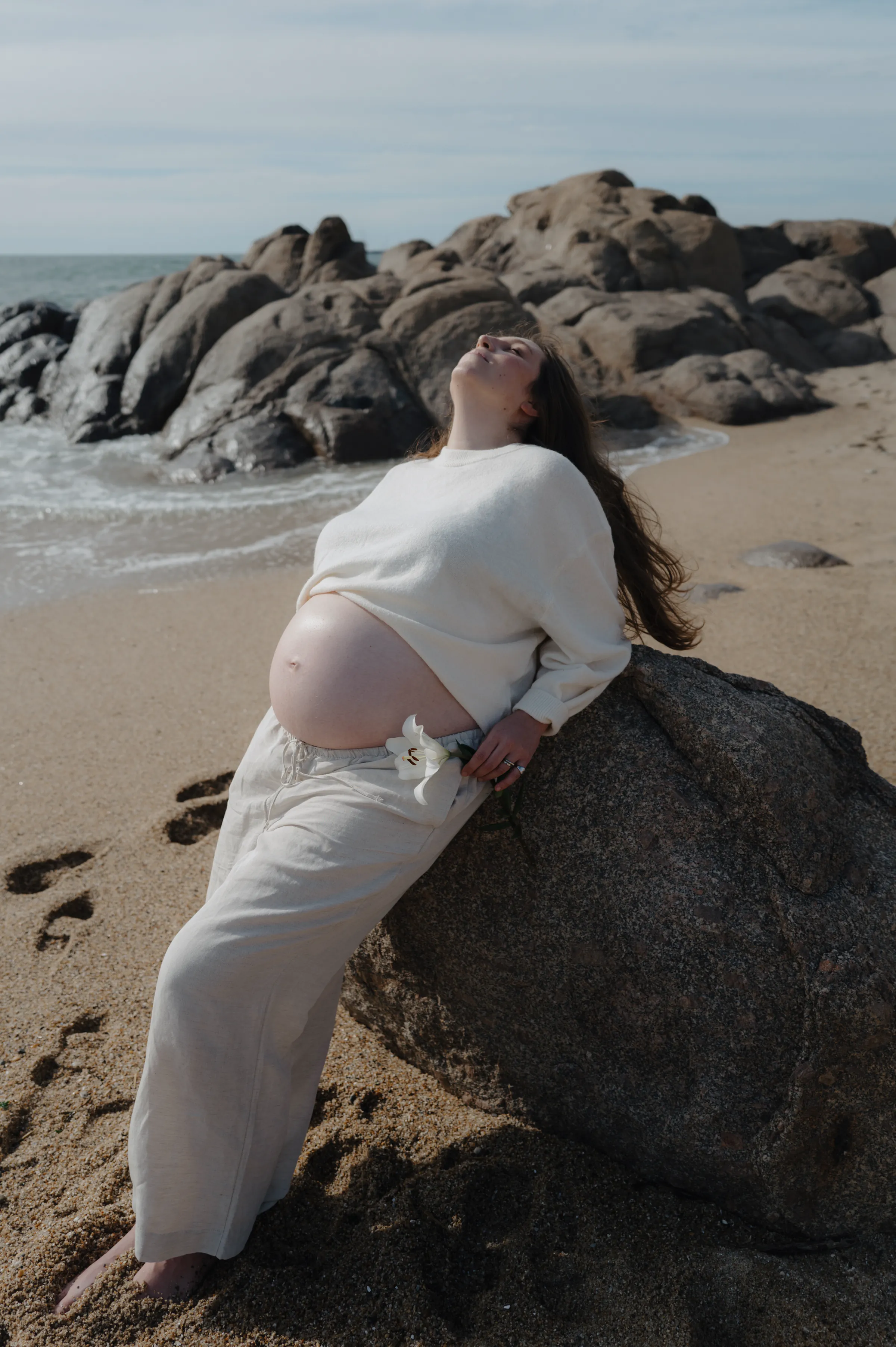 Seaside portrait of a pregnant adult reclining against a rock in soft light.