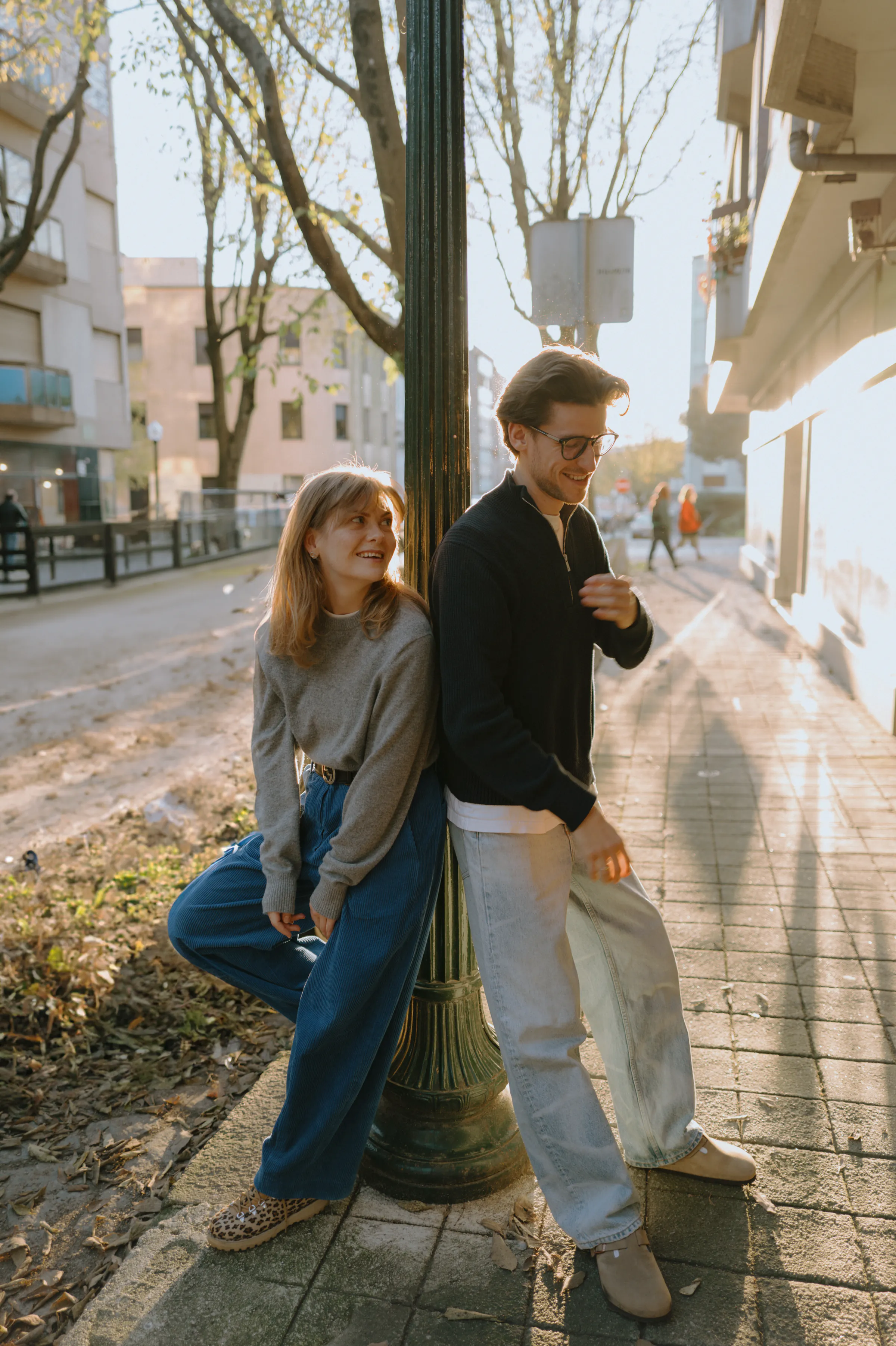 Couple during a sunny city walk, leaning by a lamppost on a quiet sidewalk in soft light.