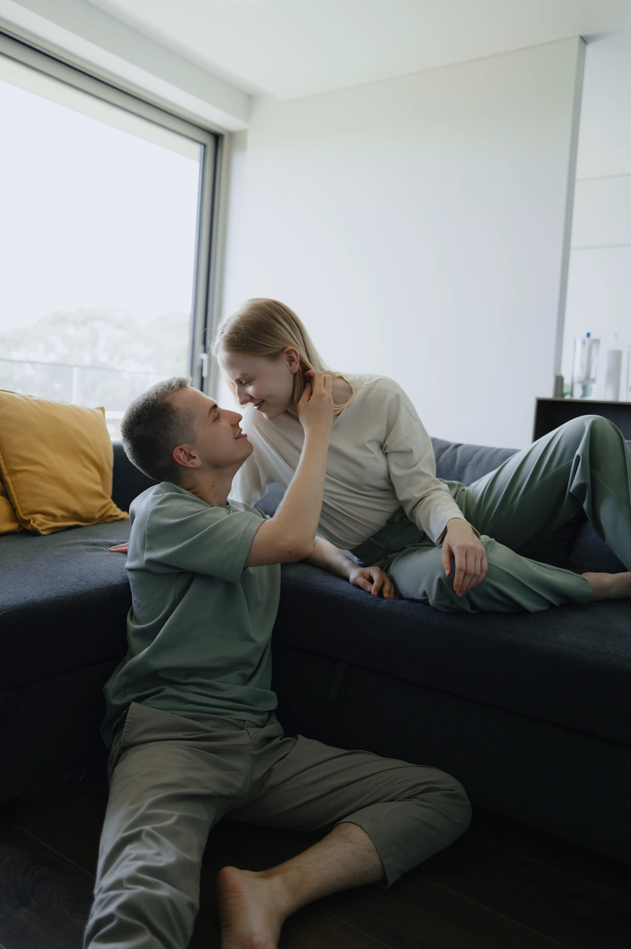 Couple in a soft light home session, sitting close together by a sofa near a large window.