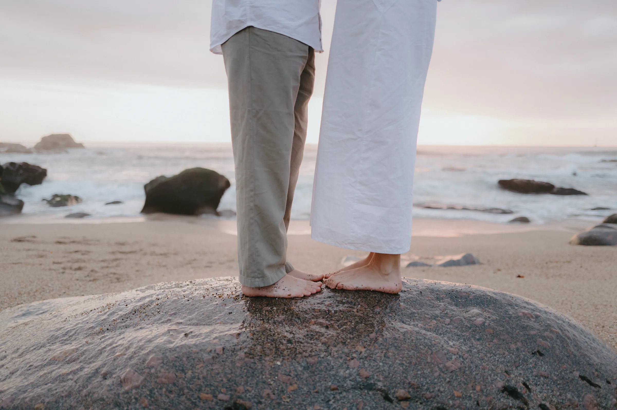 Couple standing barefoot on a seaside rock during a soft light outdoor session.