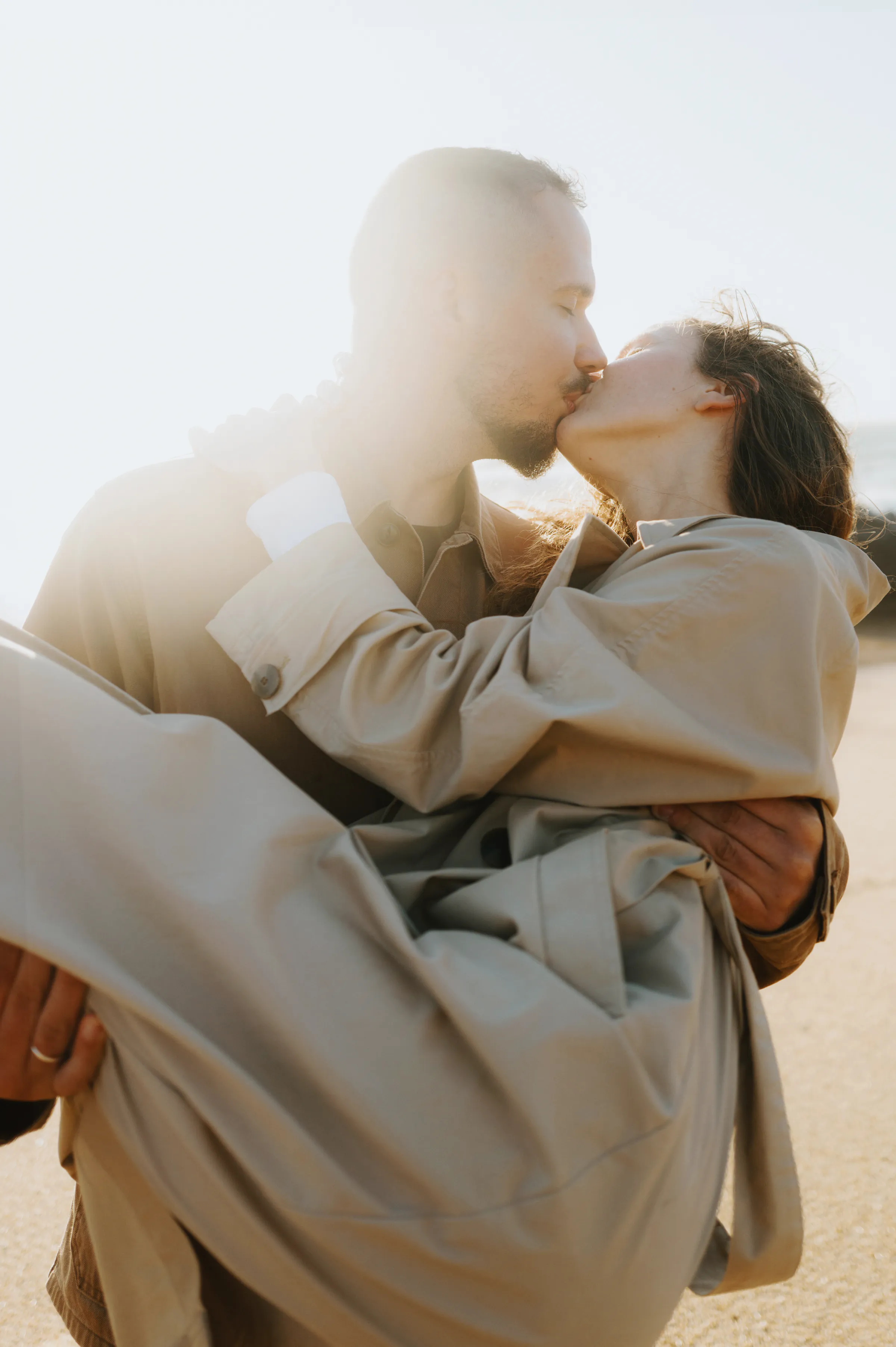 Couple kissing as one carries the other during a bright outdoor session in soft backlight.