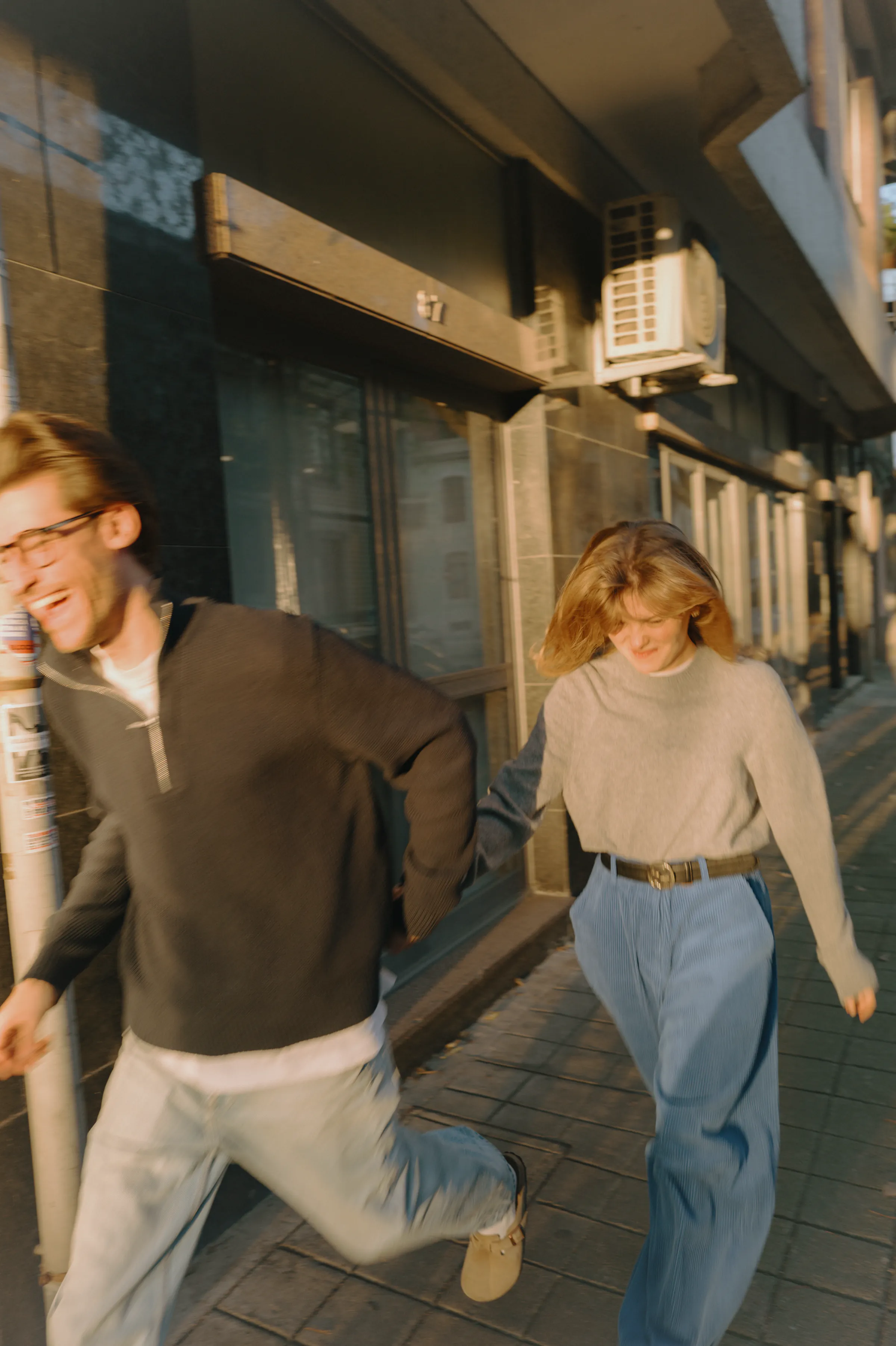 Couple during a city walk, moving along a sunlit sidewalk outside storefronts in soft light