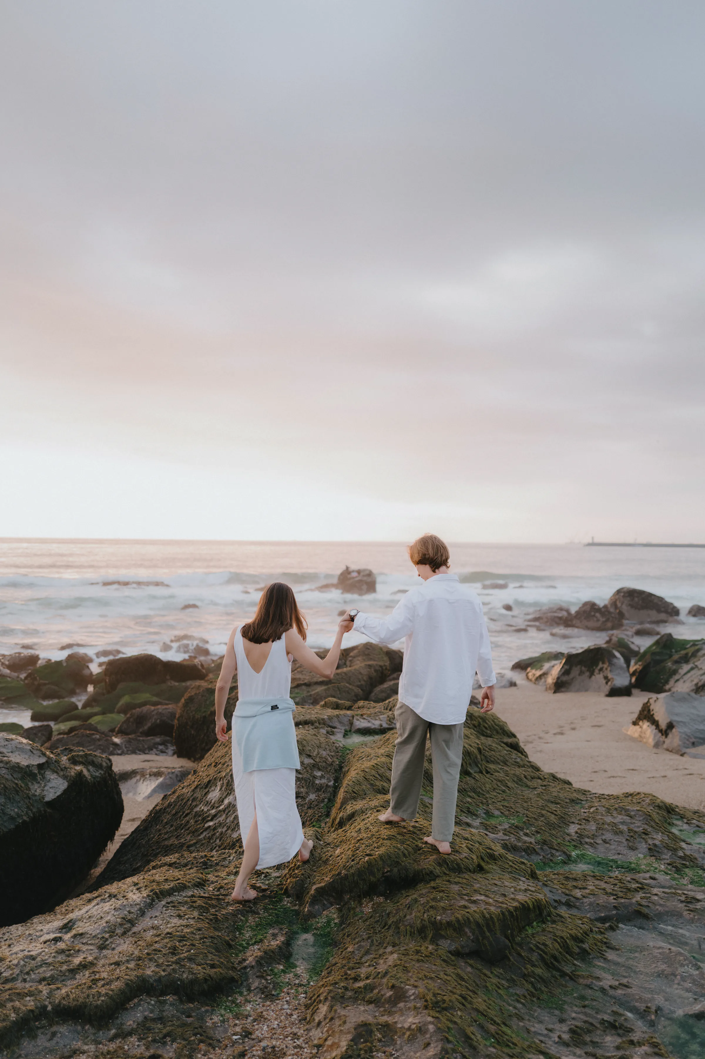 Couple walking hand in hand across seaside rocks during a soft light outdoor session.