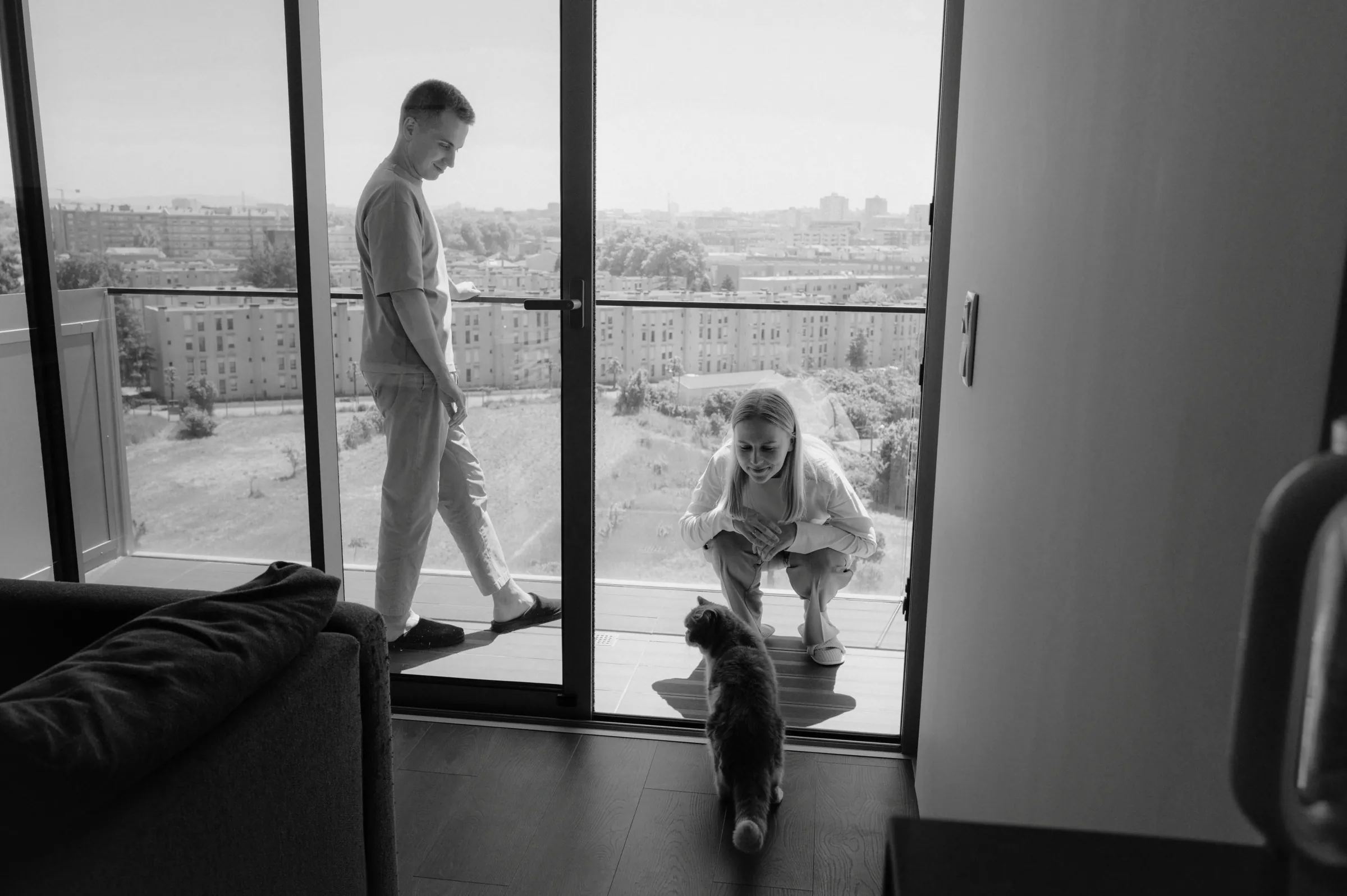 Home session with two adults by balcony doors, a small dog, and a soft city view beyond.