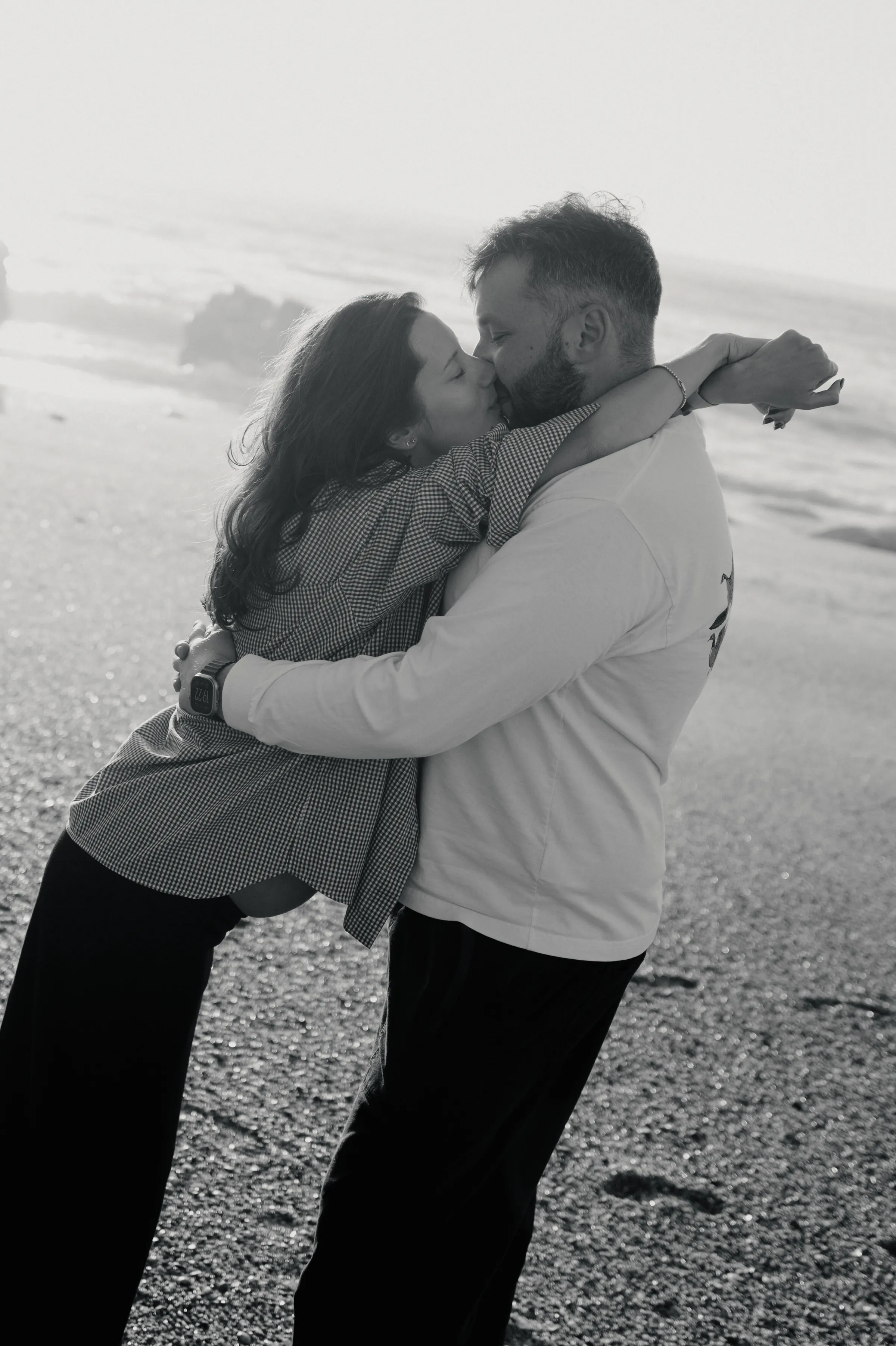 Couple embracing and kissing during a black-and-white seaside outdoor session on a pebbled beach.