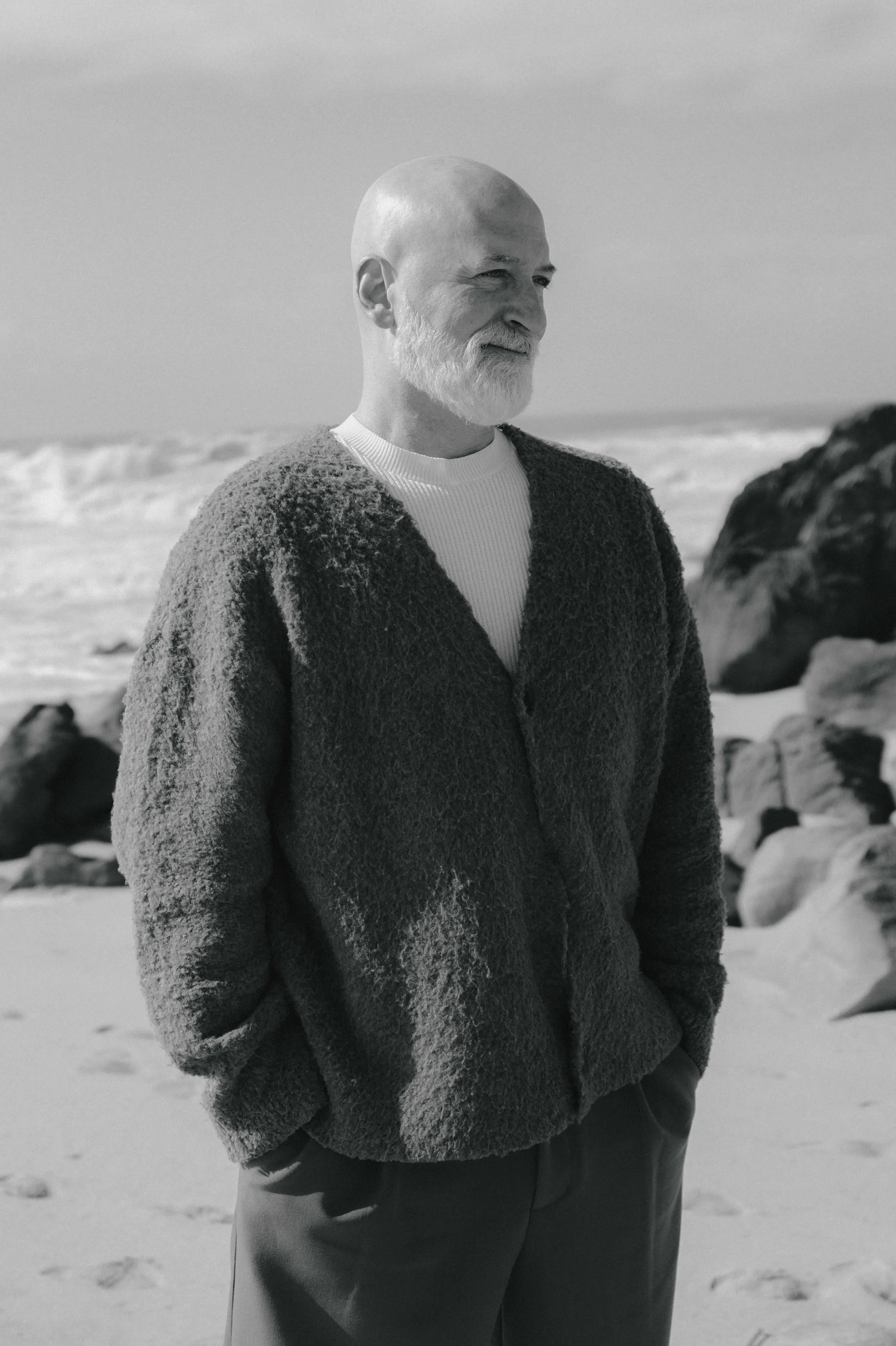 Black-and-white seaside portrait of a bearded man in a textured cardigan standing on a rocky beach.