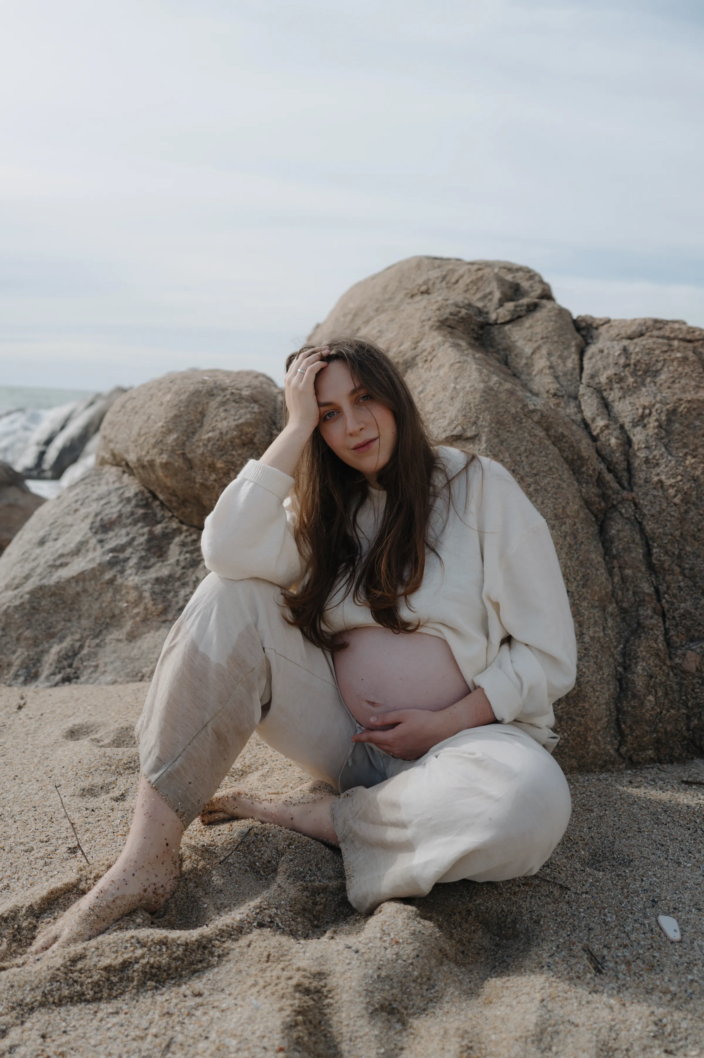 Outdoor seaside portrait of a seated woman in light clothing among rocks and sand.