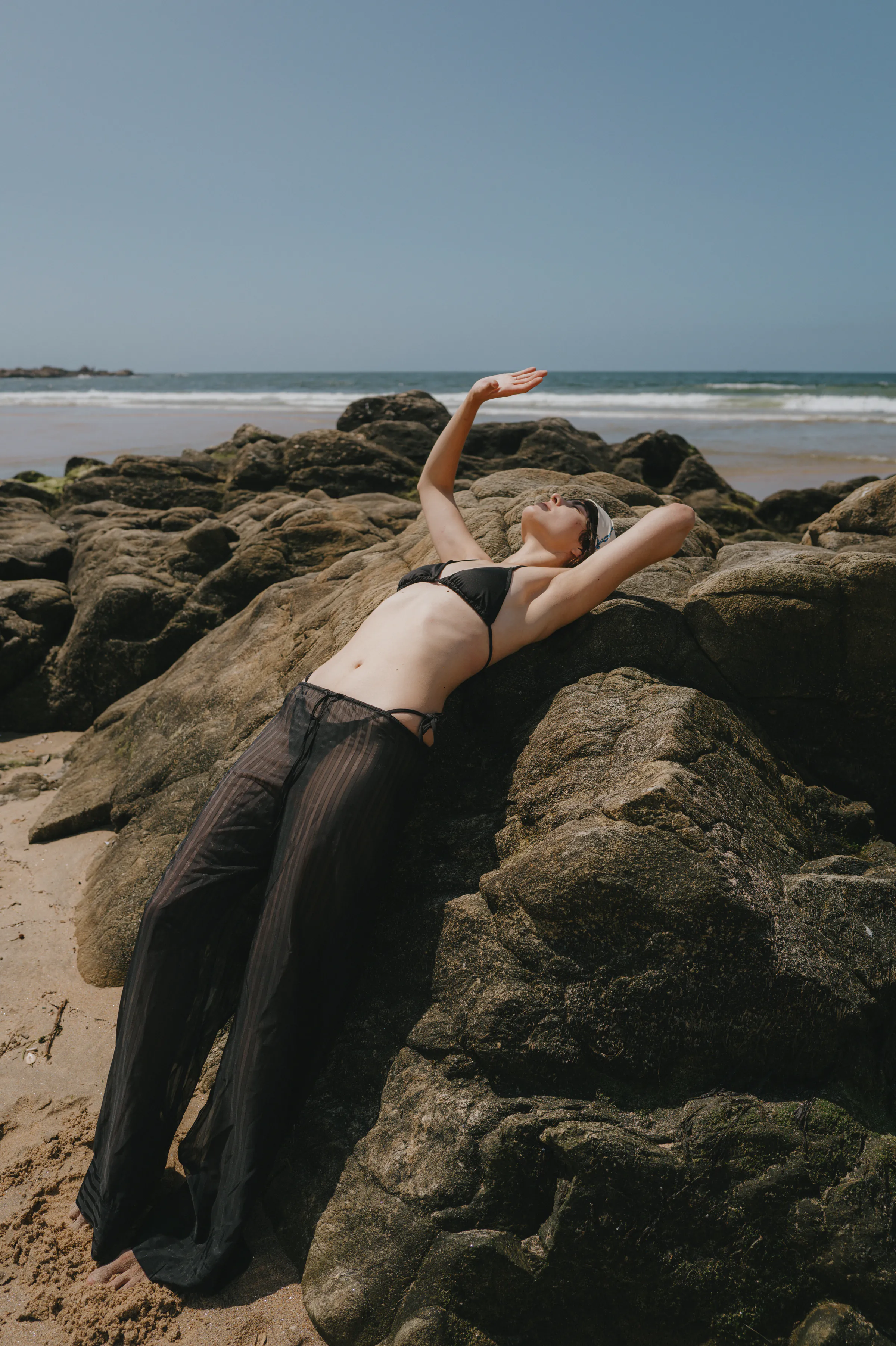 Seaside portrait of an adult reclining on sunlit rocks, with waves and open sky in the background.
