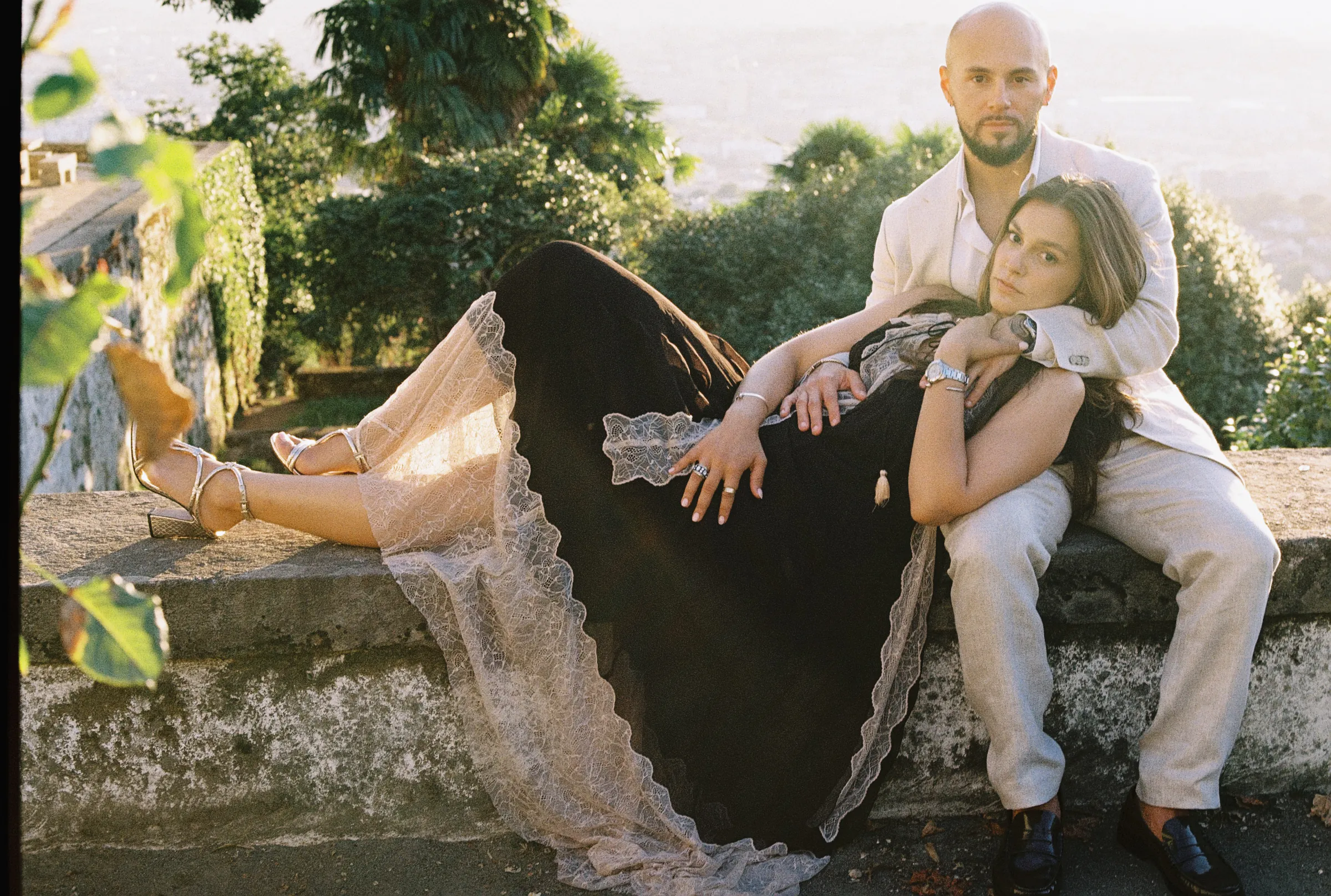 Couple portrait on a stone wall during an outdoor session with greenery and water in soft light.