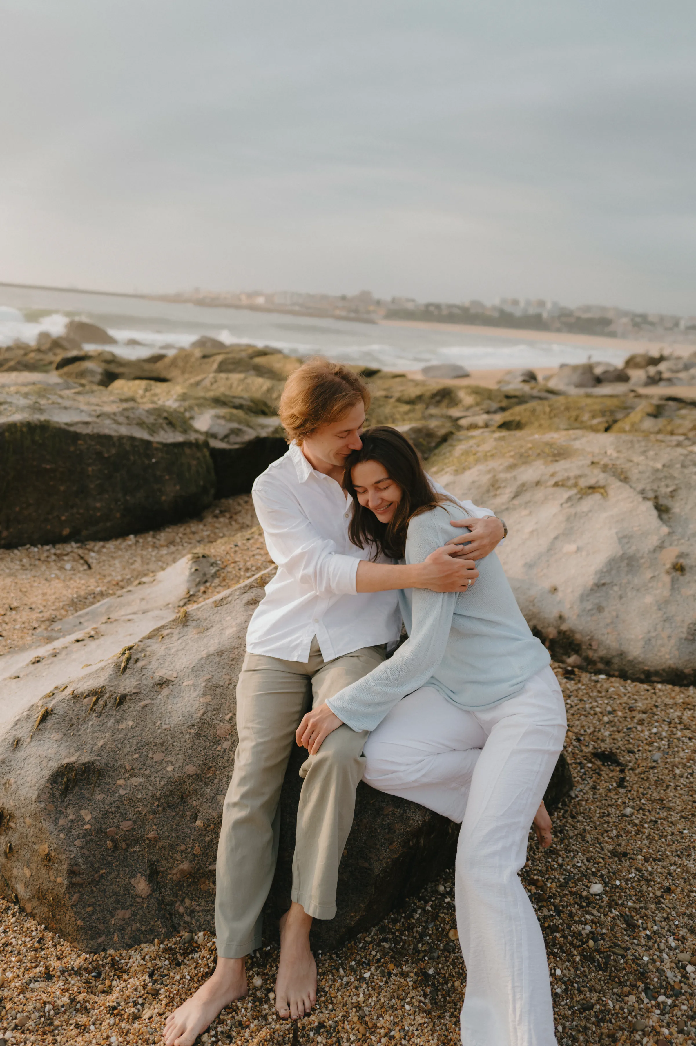 Couple portrait on seaside rocks, sitting close together in soft light with waves behind them.