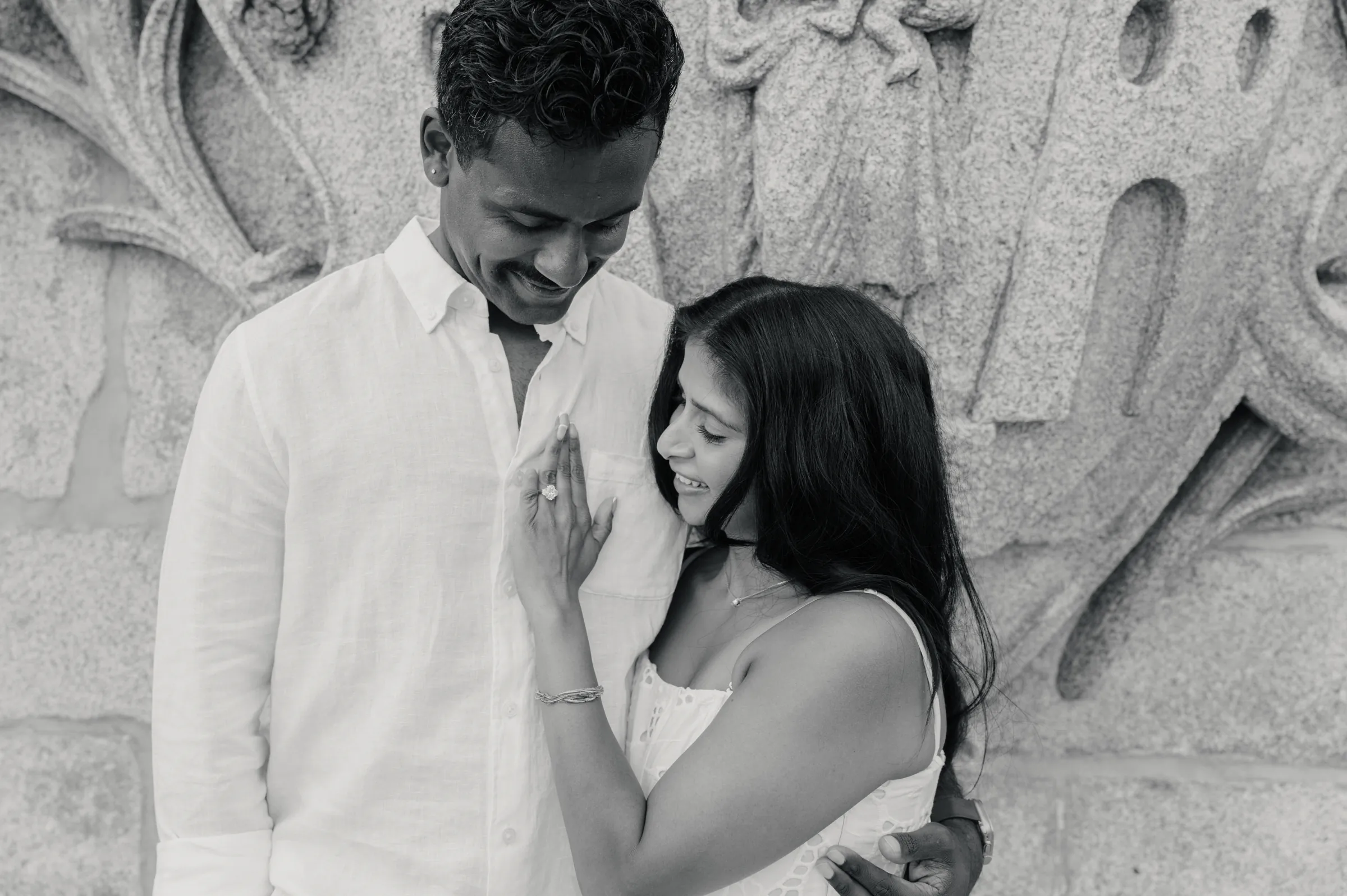 Black-and-white couple portrait beside a textured stone wall, standing close in soft light.