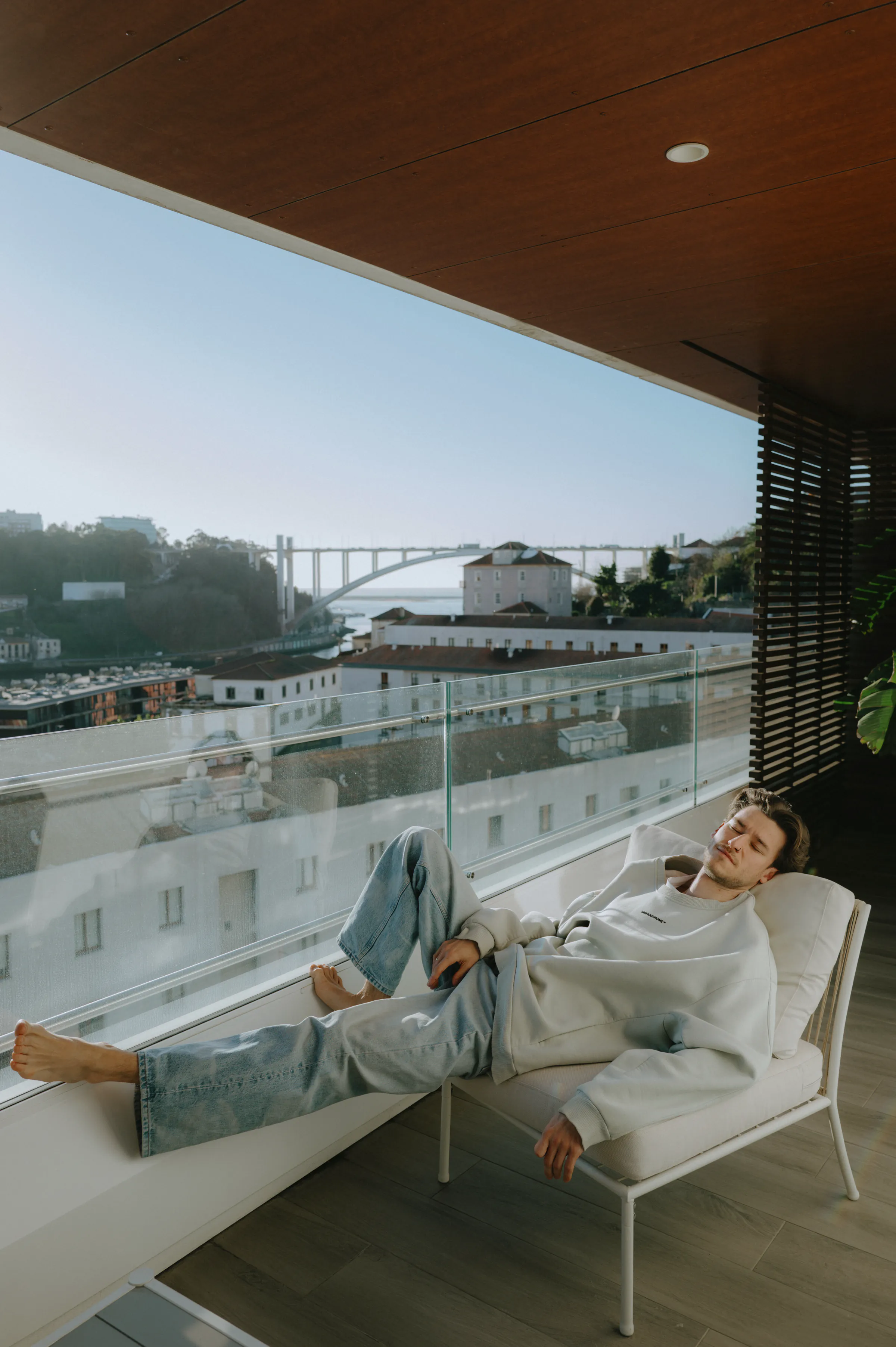Balcony portrait of a person reclining on a lounge chair overlooking a river, bridge, and city skyline.