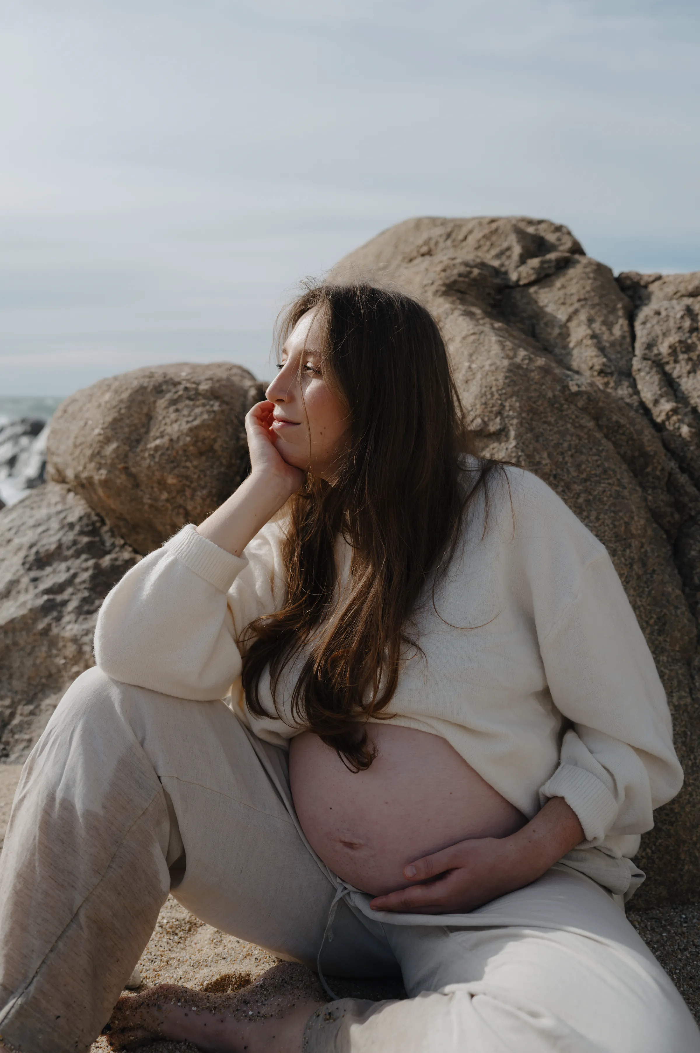 Maternity portrait on seaside rocks, seated in soft light with one hand resting on belly.