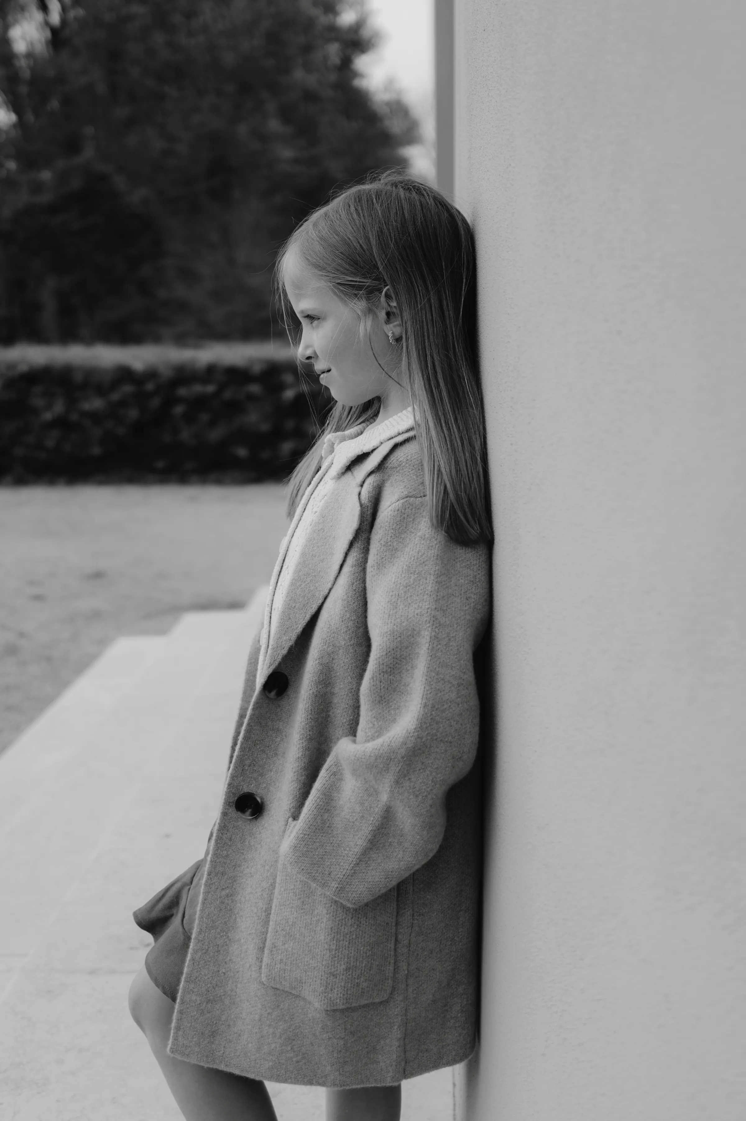 Black-and-white outdoor child portrait, leaning against a wall in a long coat with soft background blur.