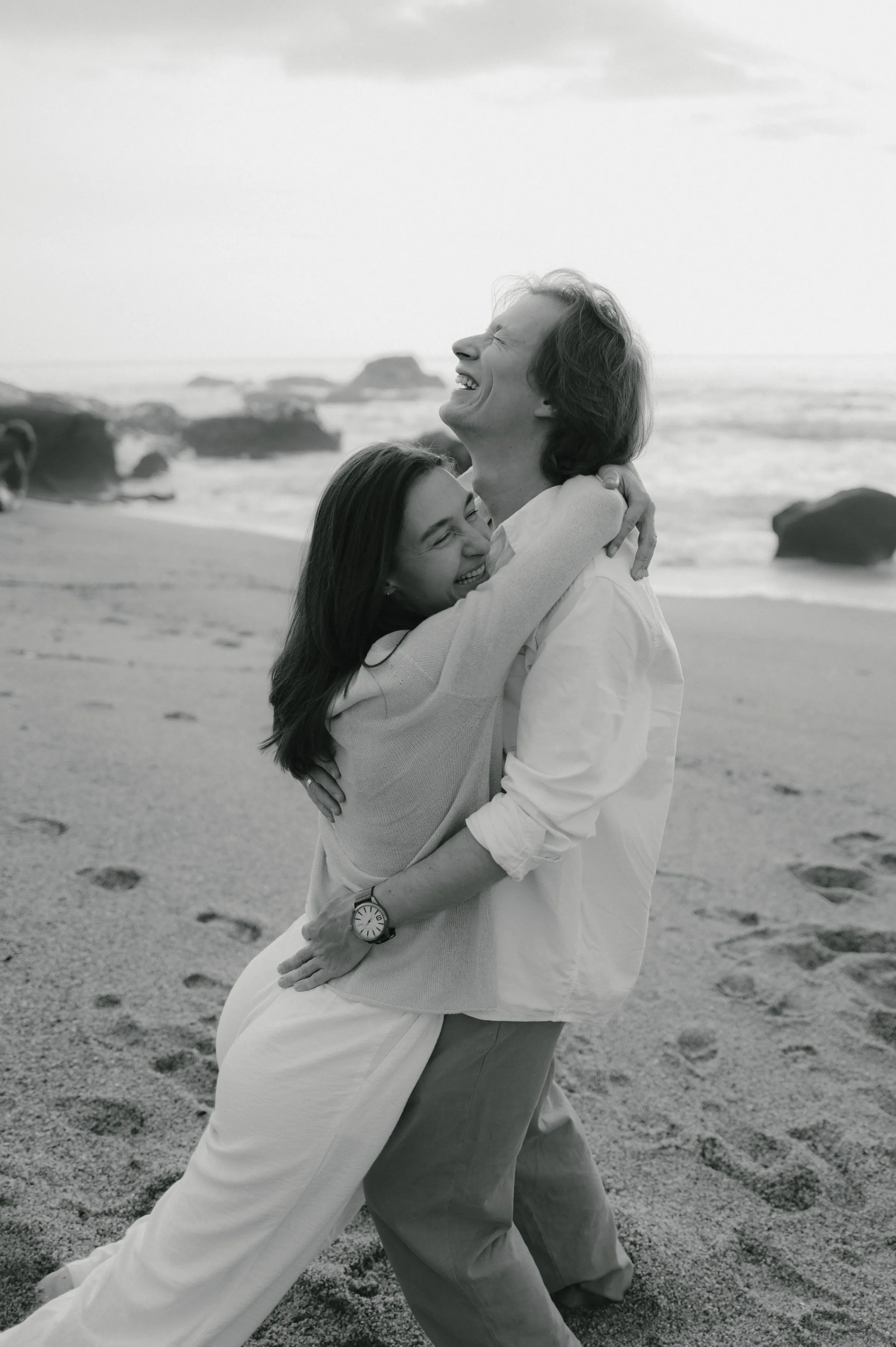 Black-and-white seaside portrait of a couple embracing on a sandy beach in soft coastal light.