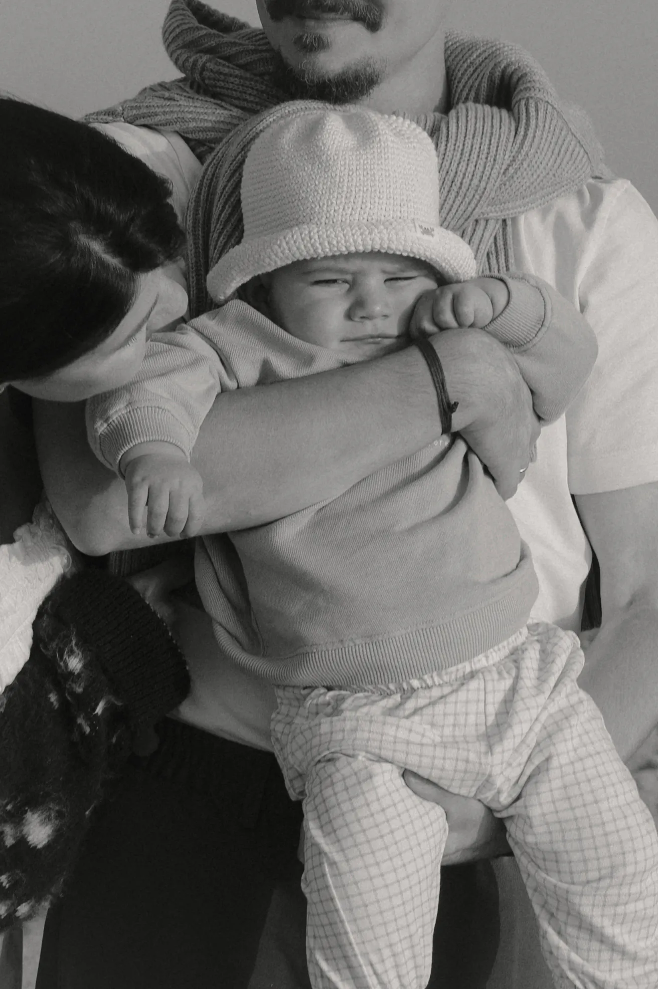 Black-and-white close portrait of a family holding a baby in a knit hat, softly framed indoors.