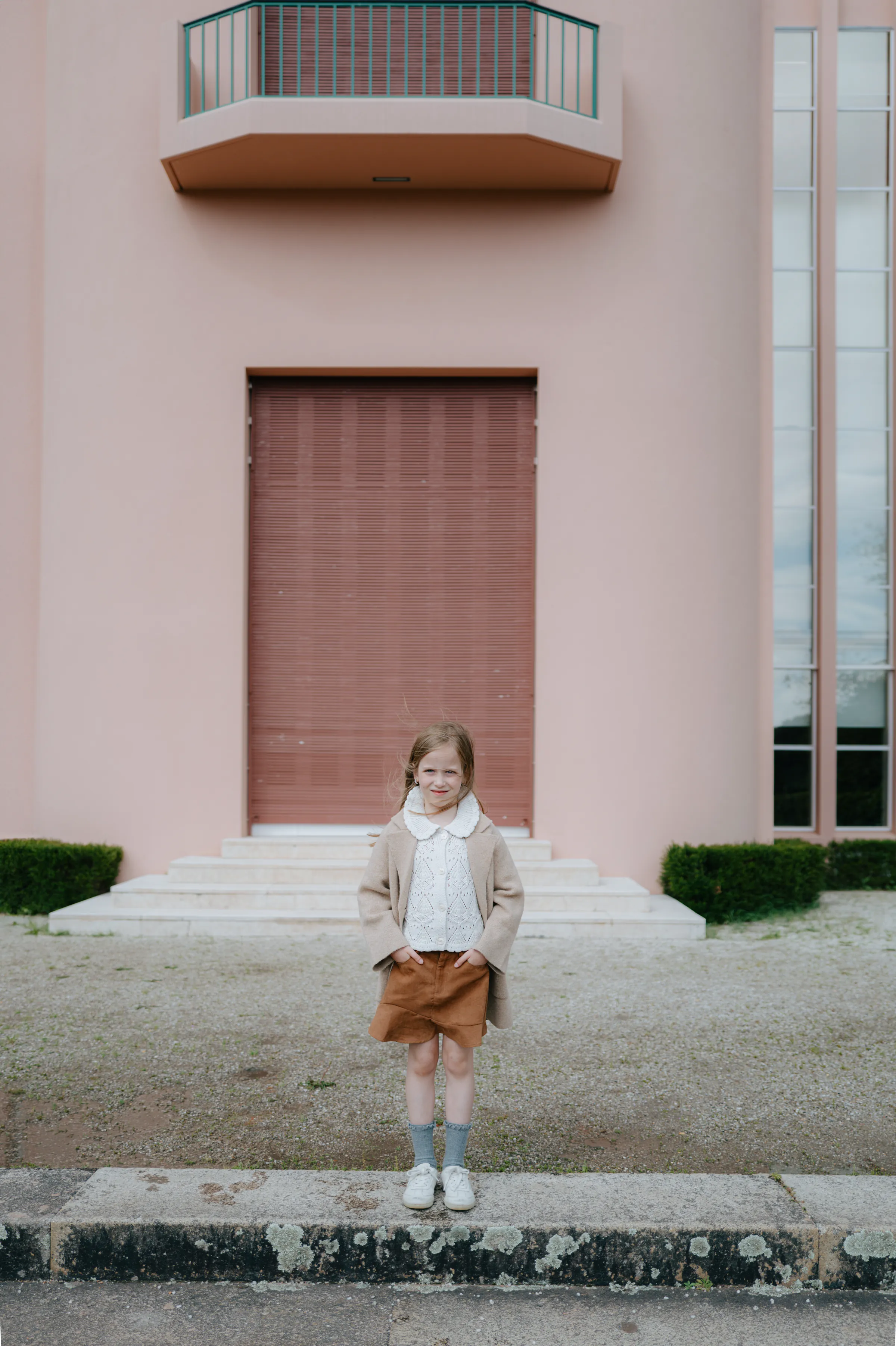 Child portrait during an outdoor session, standing by a pink building facade with soft light and simple architectural lines.