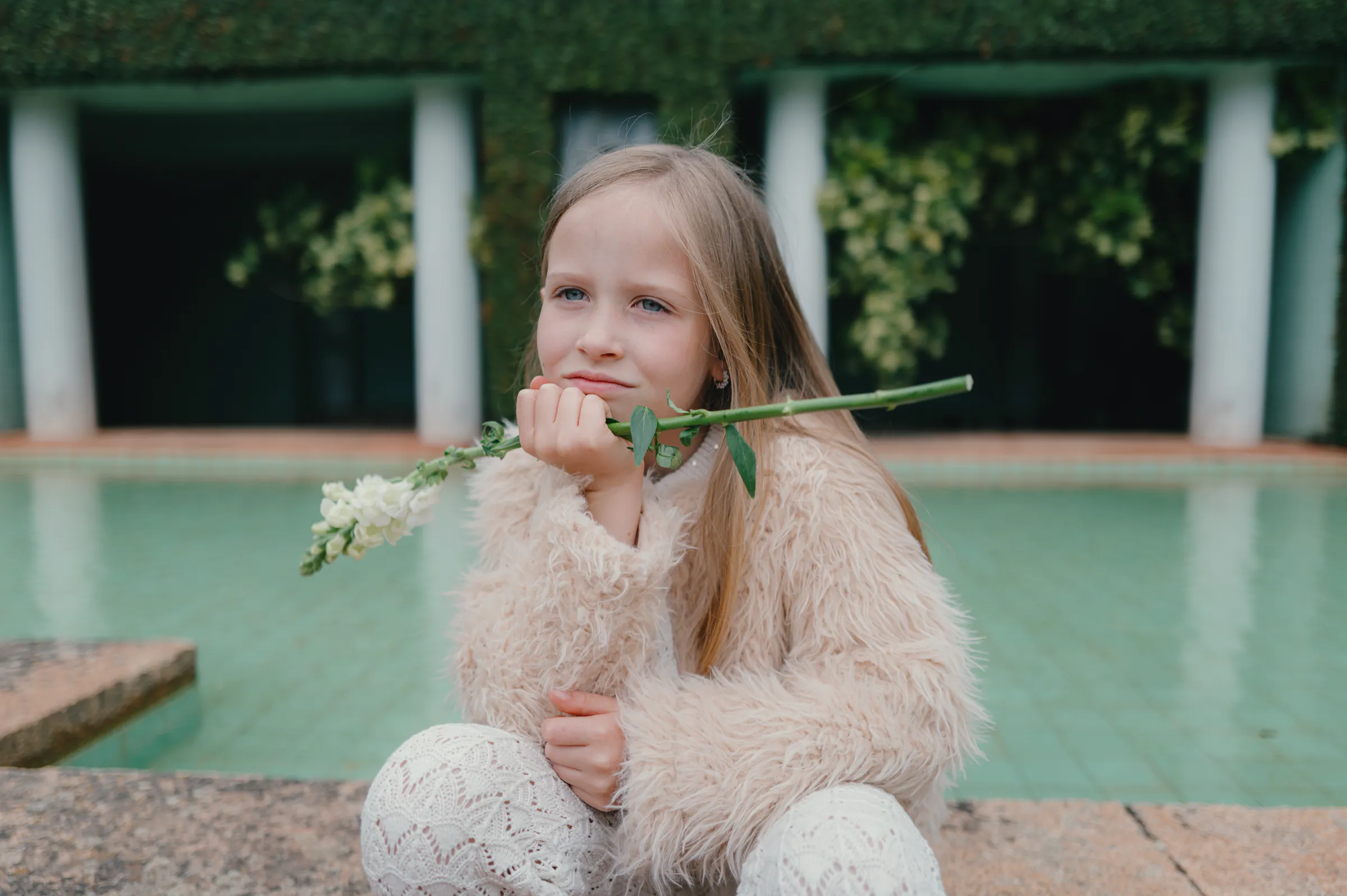 Outdoor child portrait by a pool, holding a long-stemmed flower in soft garden light.