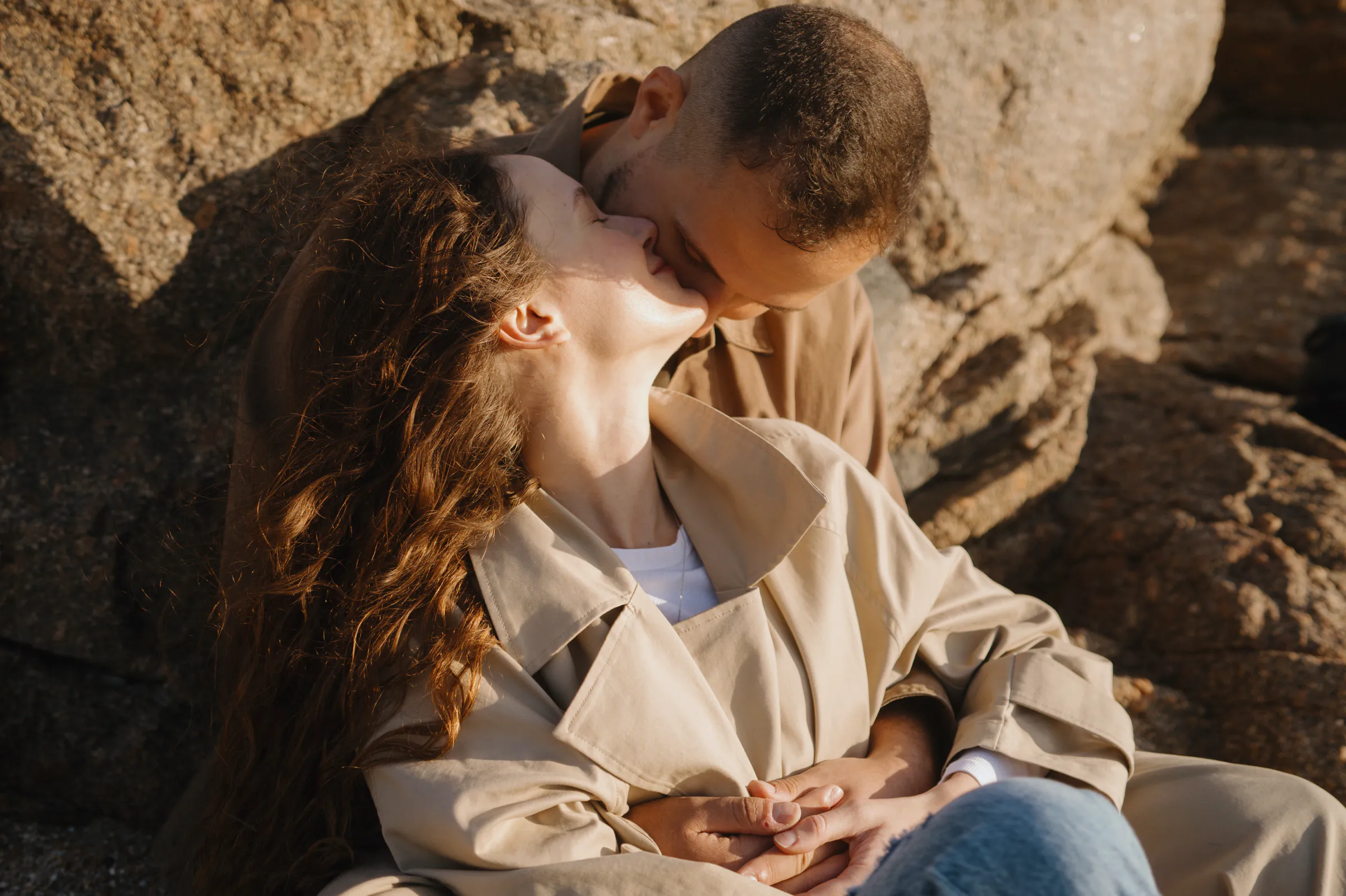 Couple kissing during an outdoor session against sunlit rocks, framed as a close portrait in soft light.