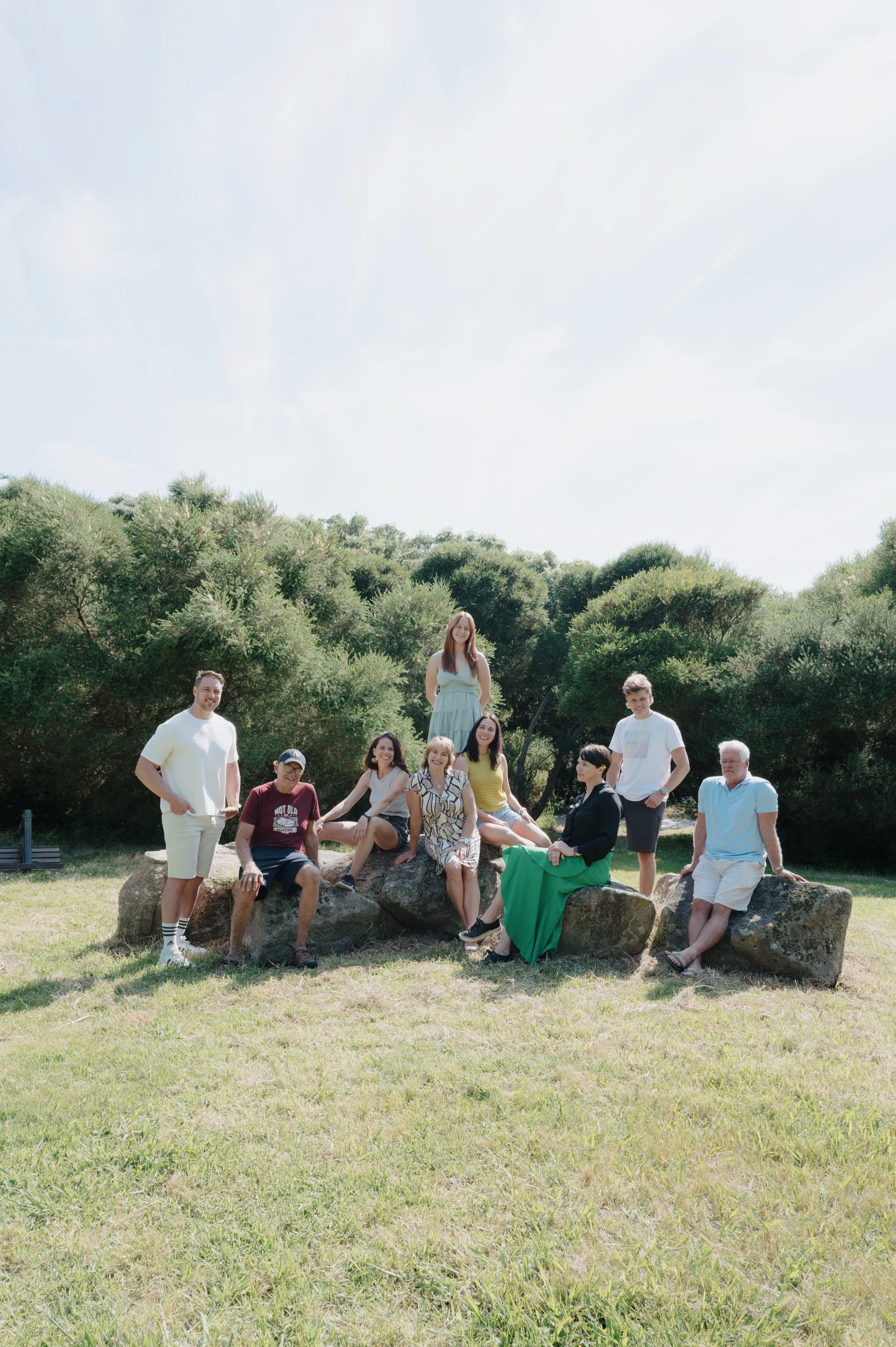 Family outdoor session in soft light, gathered on rocks in a grassy park with trees behind them.