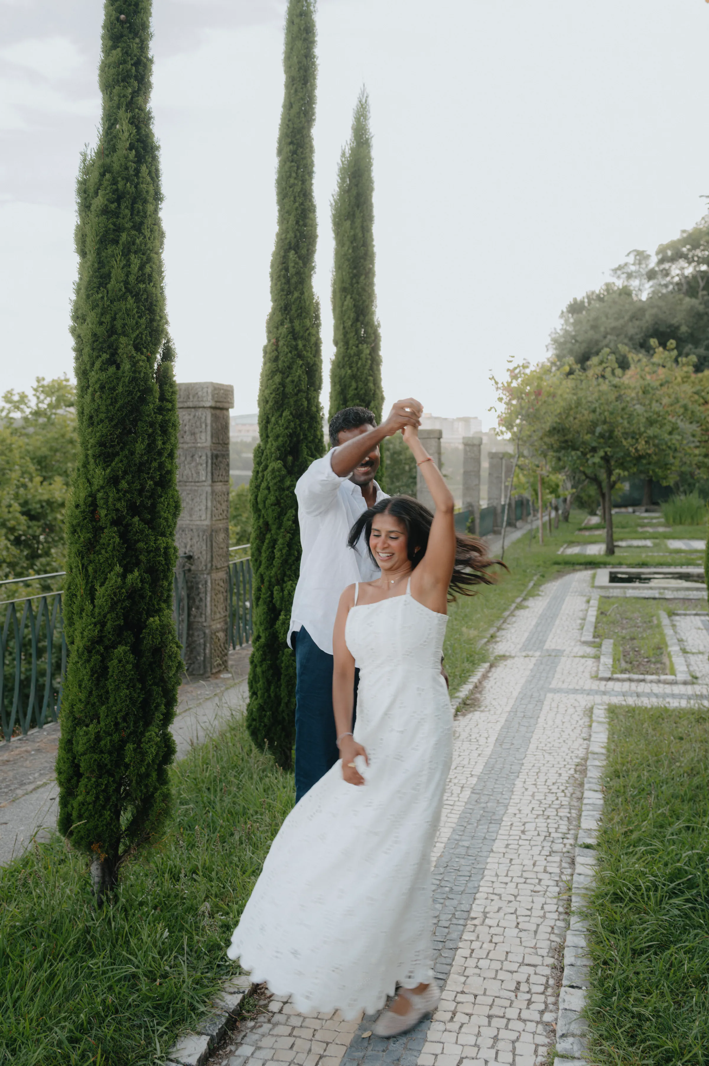 Couple twirling on a garden path lined with tall cypress trees during an outdoor session.
