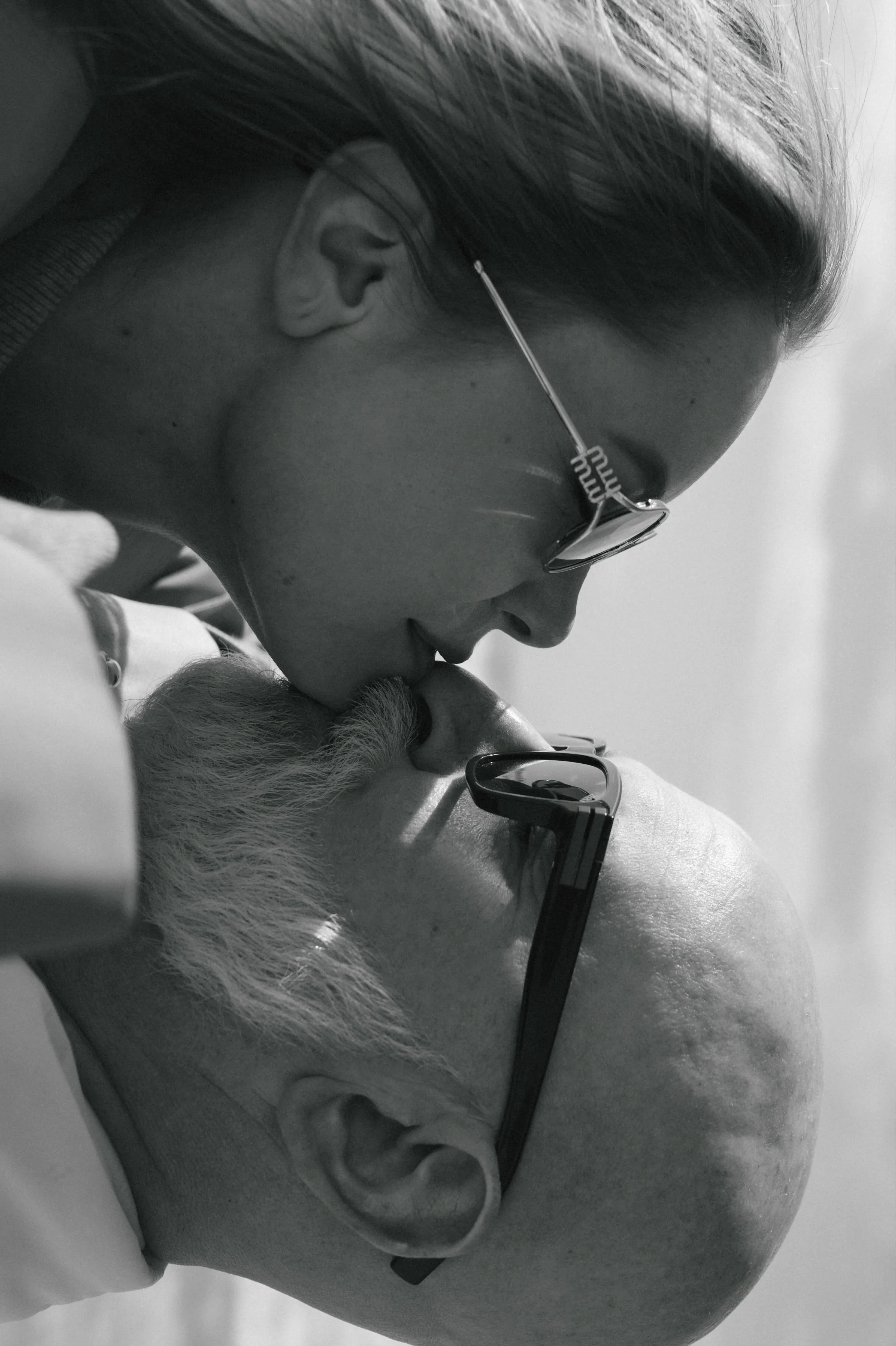 Black-and-white close portrait of a couple in sunglasses sharing a kiss in soft light.