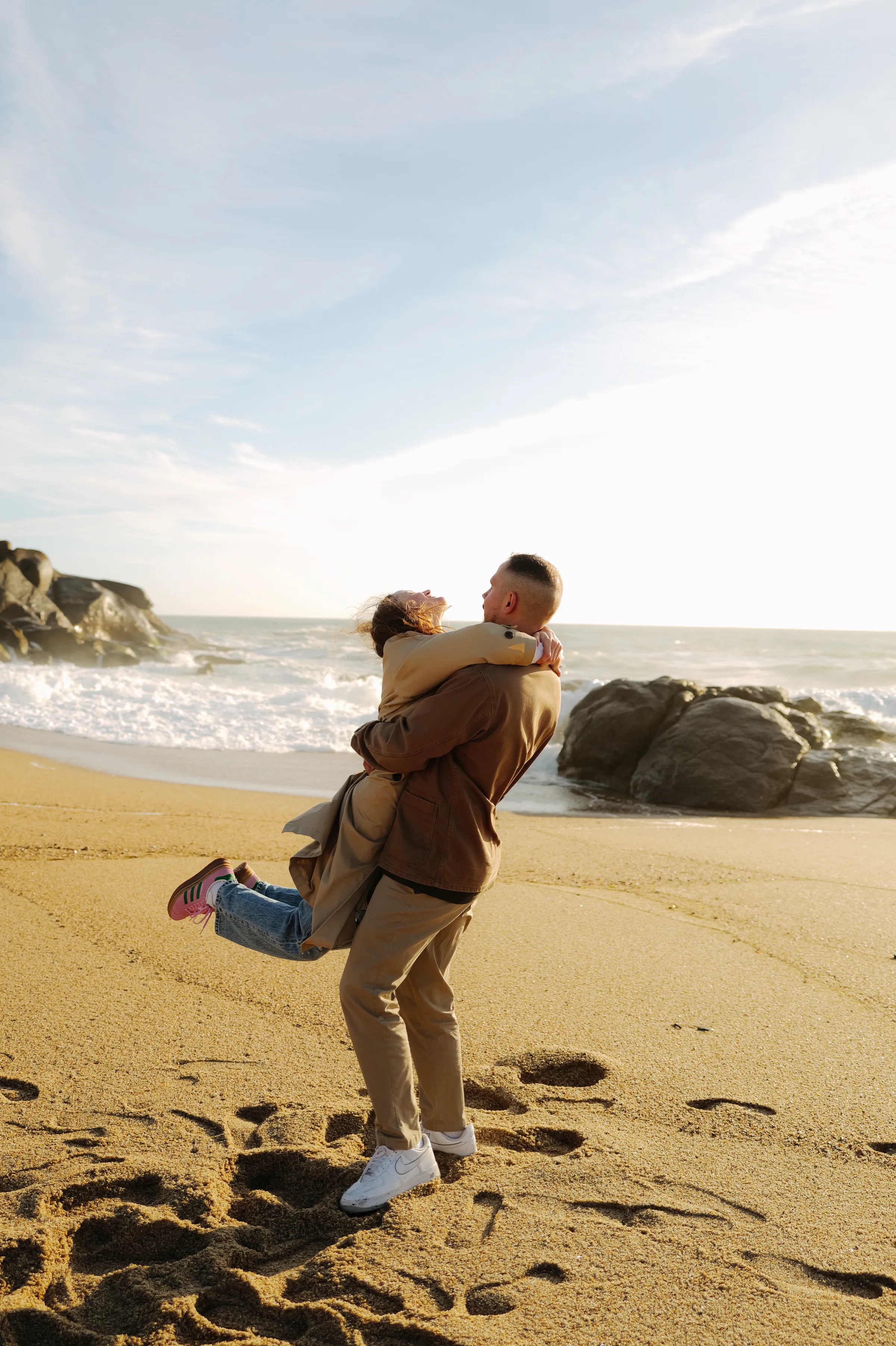 Seaside portrait of an adult lifting a child on a sandy beach beside rocks and waves.