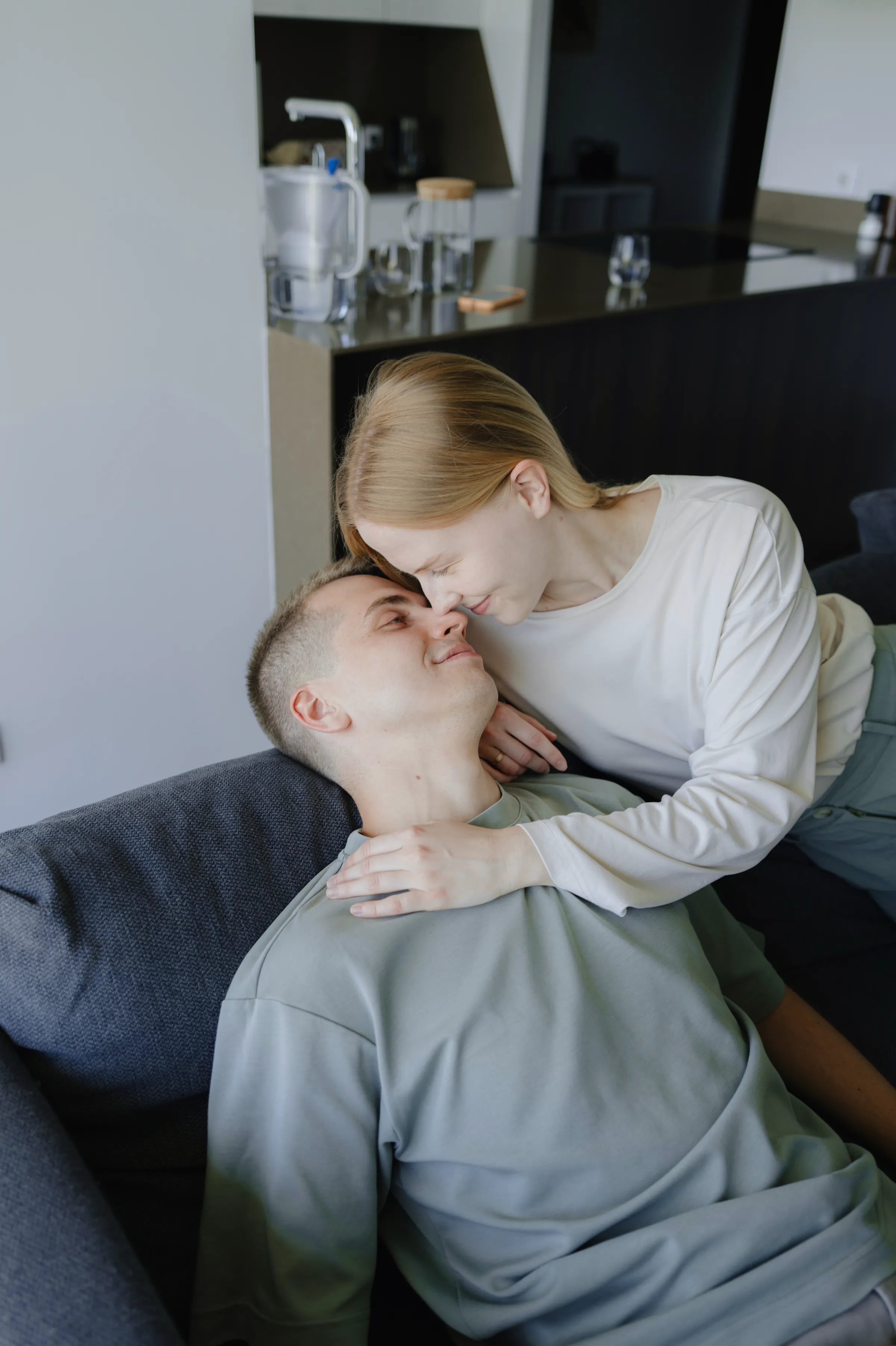 Couple close portrait on a sofa during a relaxed home session, with soft indoor light and a kitchen behind them.
