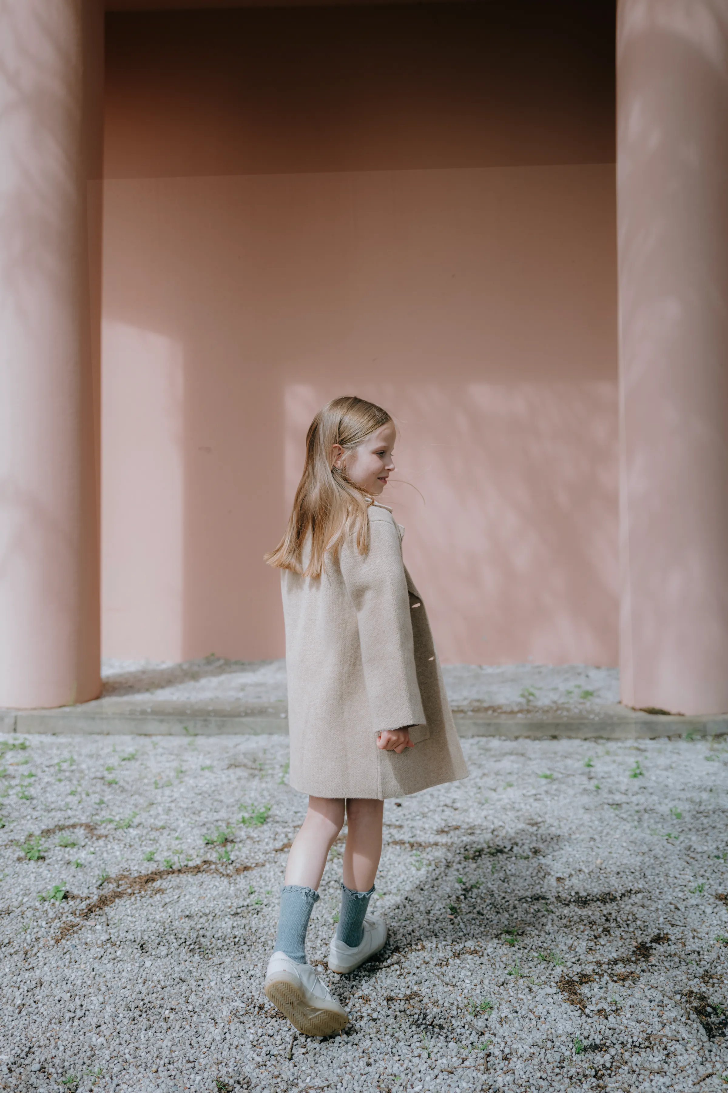 Child portrait during an outdoor session, walking through soft light between pale pink columns.
