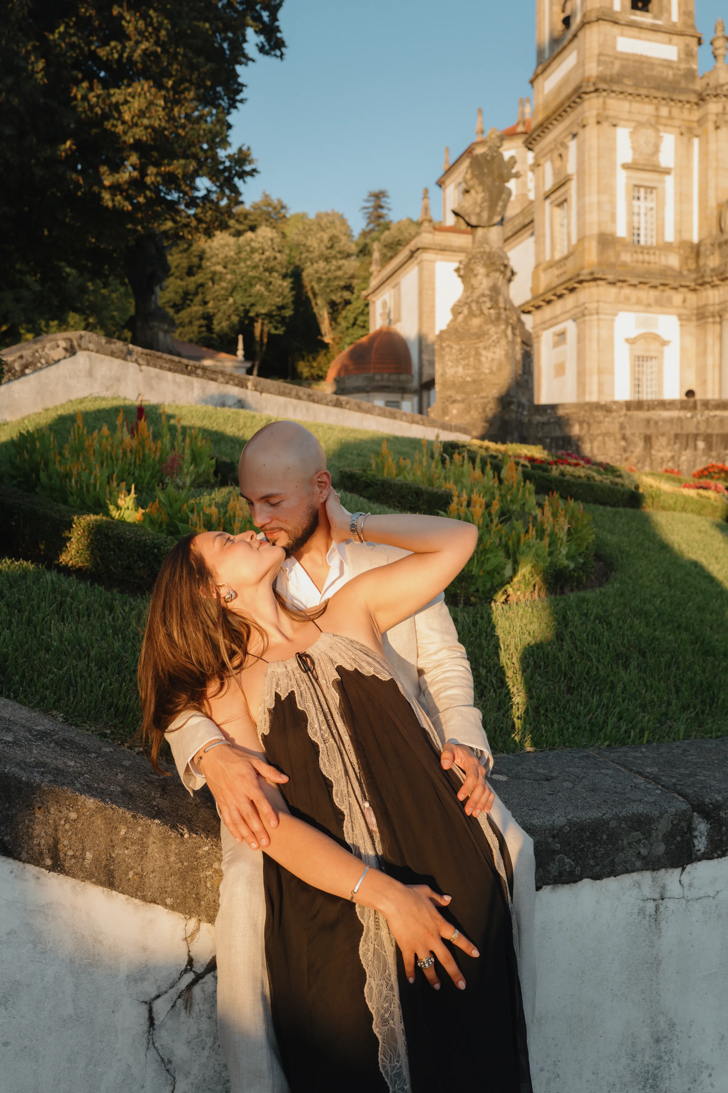 Couple portrait in a formal garden beside an ornate historic building, captured in warm soft light.