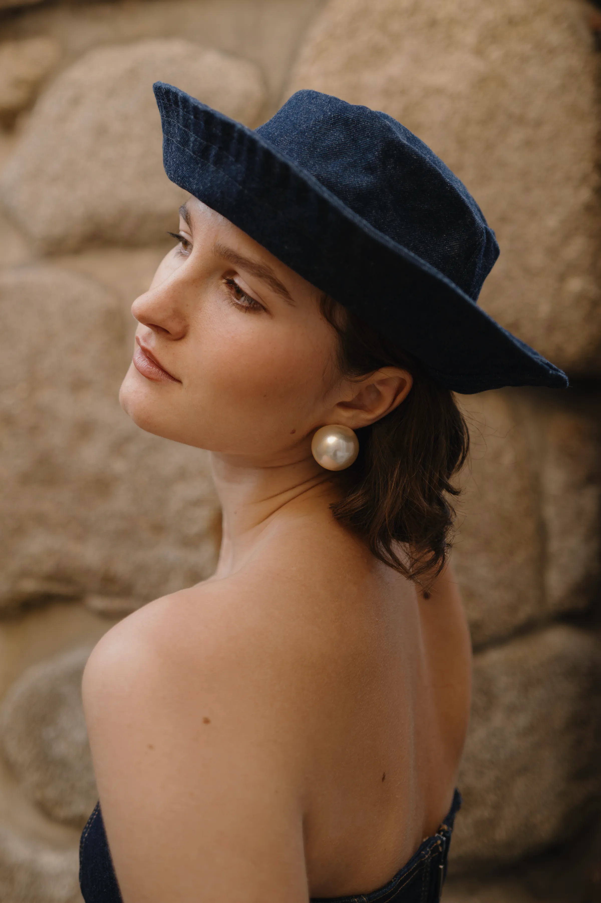 Close portrait with wide-brimmed hat and pearl earring, posed against a textured stone wall.
