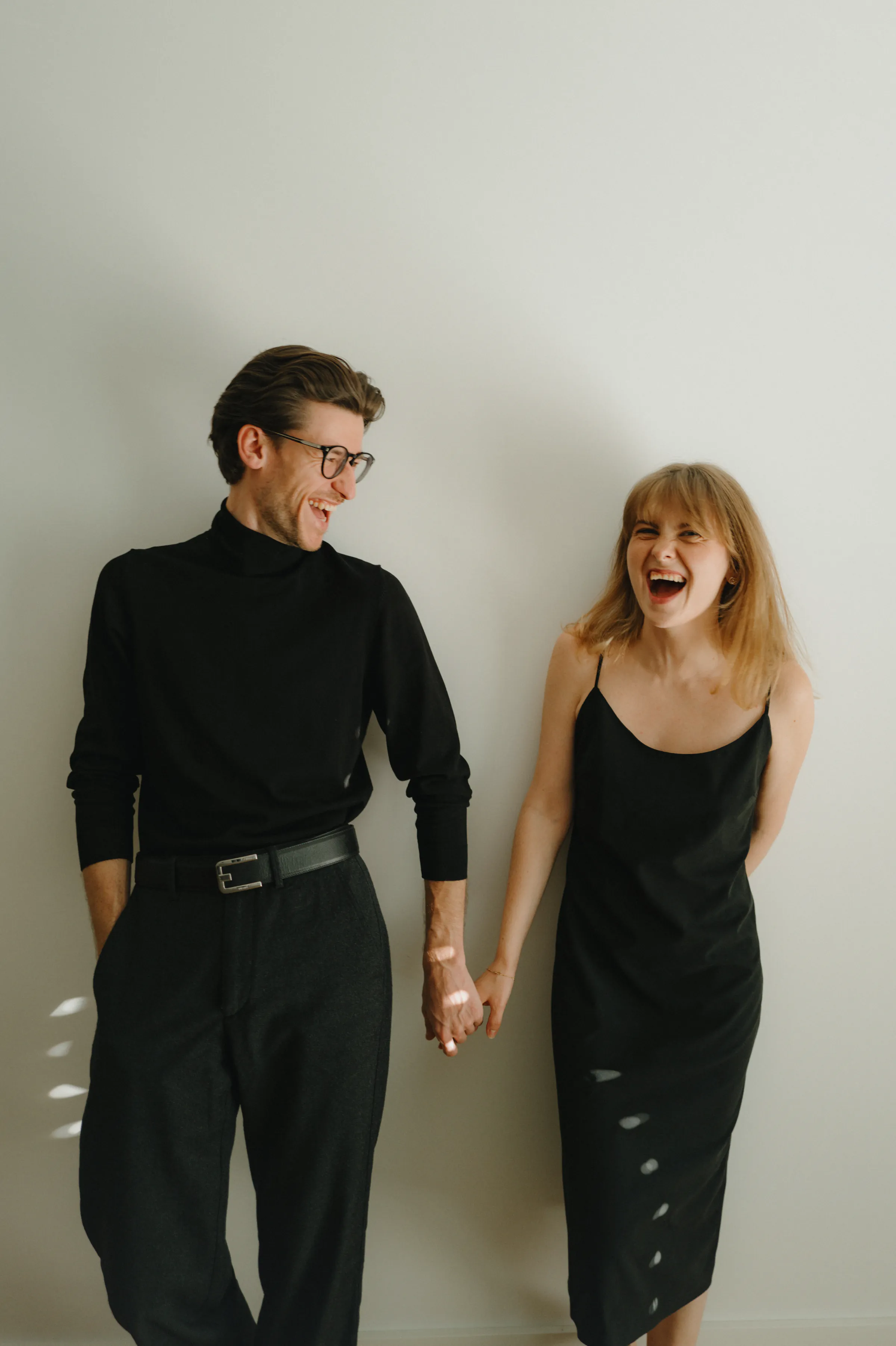 Couple portrait indoors, standing hand in hand against a plain wall in soft light.