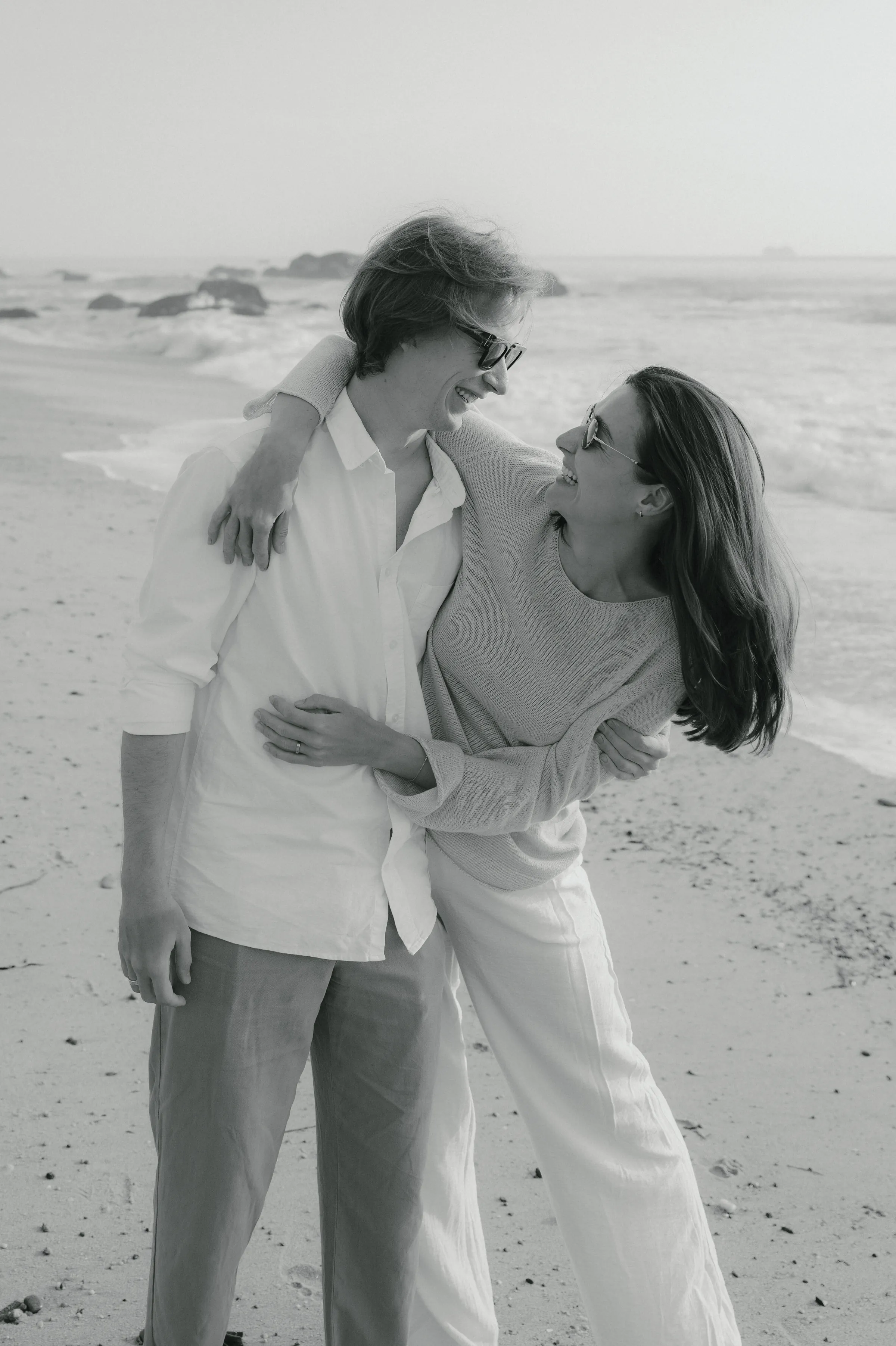 Black-and-white seaside portrait of a couple standing close together on a sandy beach in soft light.